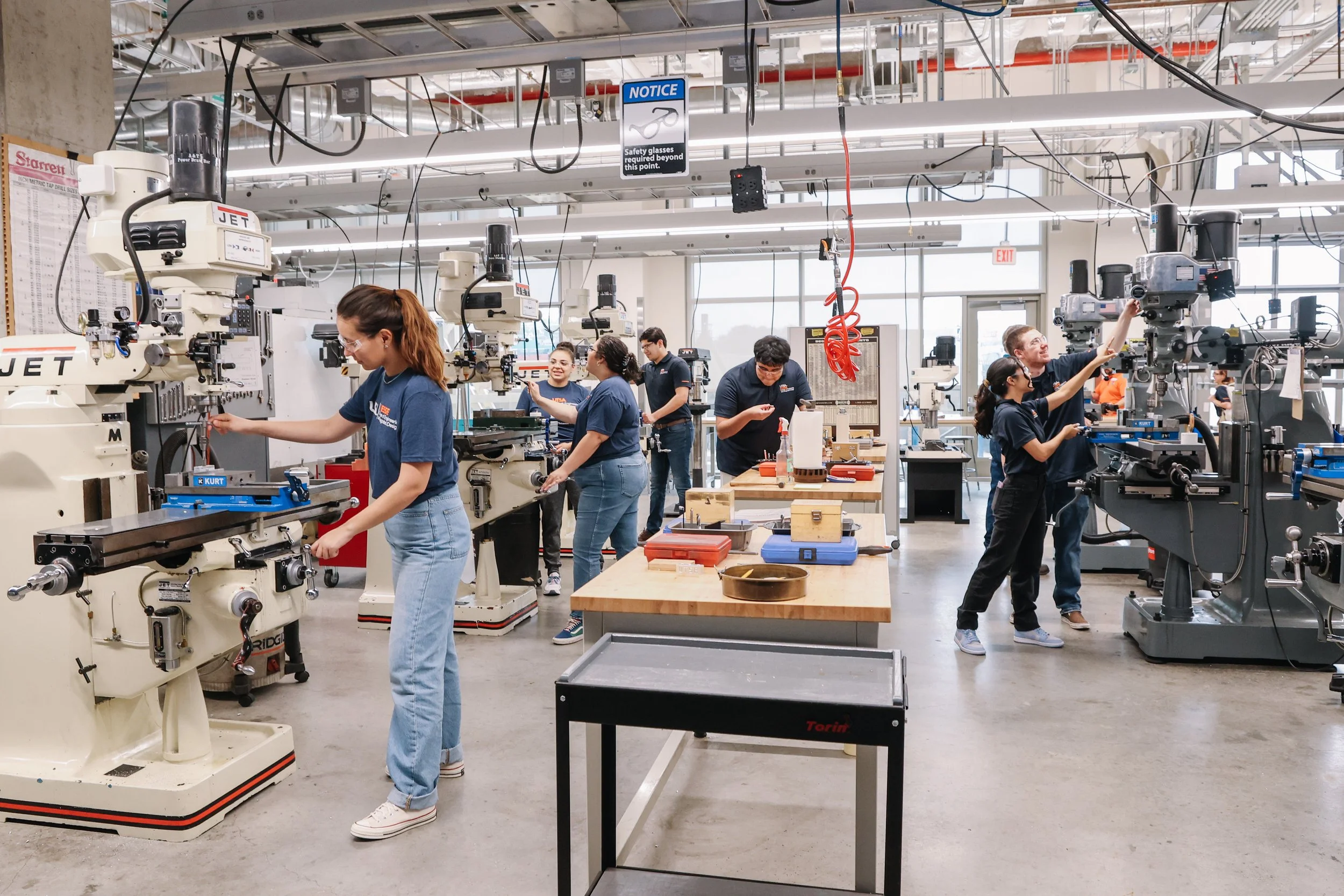 Students in a makerspace or FabLab operating machinery, with safety glasses and various tools on workbenches.
