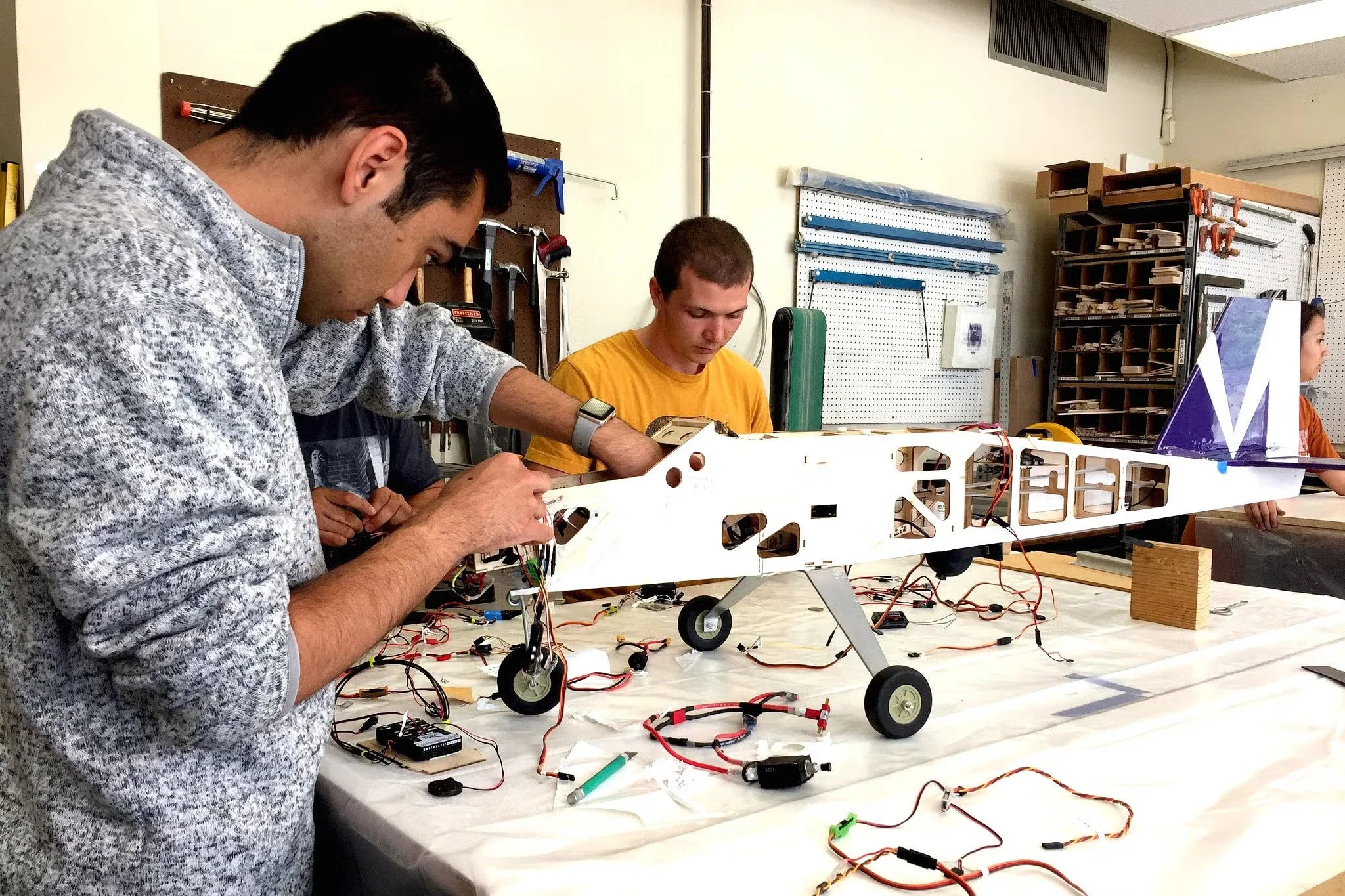Three young men working on robotics project in a workshop, with wires and parts on a table, shelves with tools and supplies in the background.