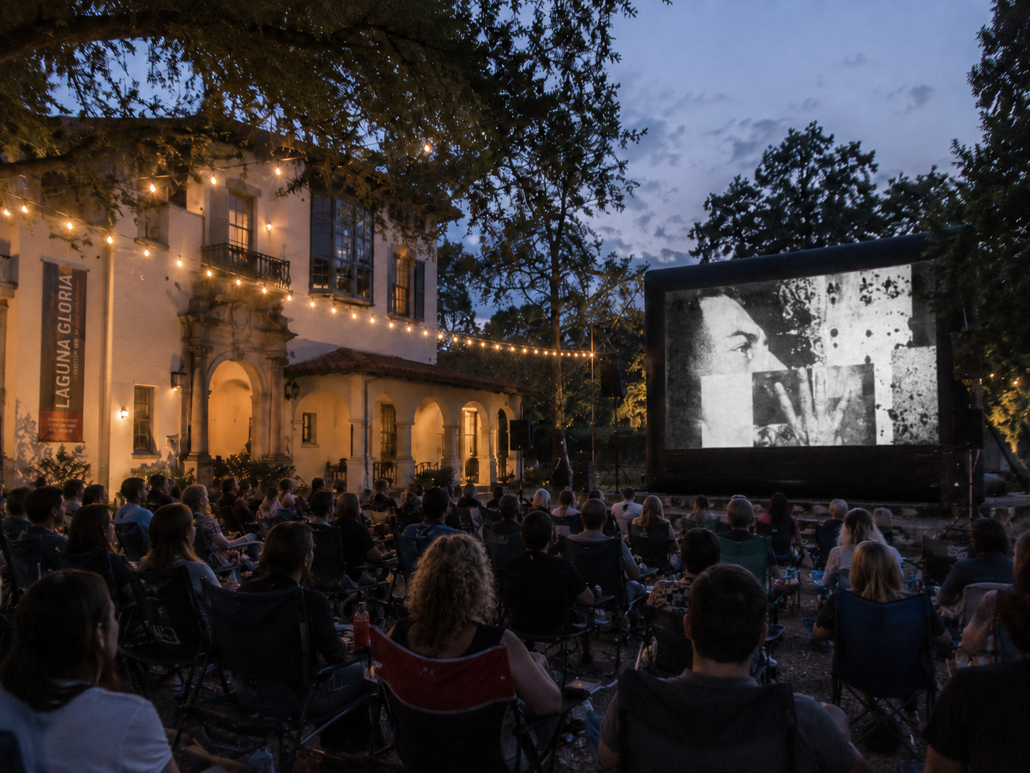 An outdoor movie screening at dusk with an audience seated in front of a large screen showing a black and white film, surrounded by string lights and a historic building with arched windows and balconies.
