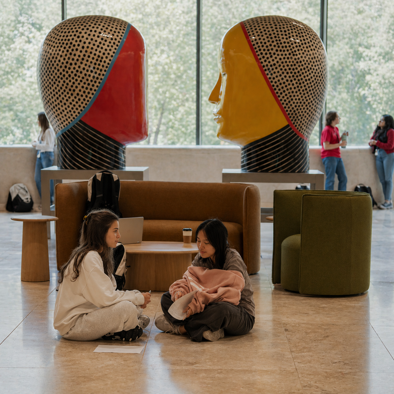 Two young medical students sitting on the floor engaged in conversation in a spacious indoor area with large art sculptures of large human heads in profile behind them and people walking in the background.