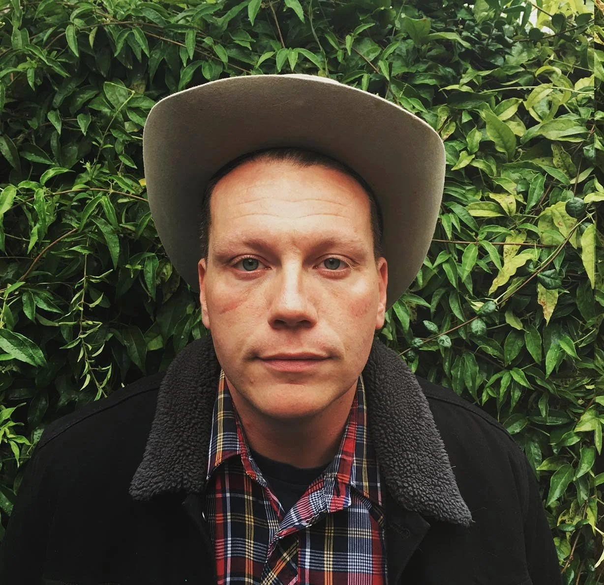 A man with light skin, short brown hair, and blue eyes wearing a tan cowboy hat, a plaid shirt, and a black jacket with a faux shearling collar, standing in front of green leafy bushes.