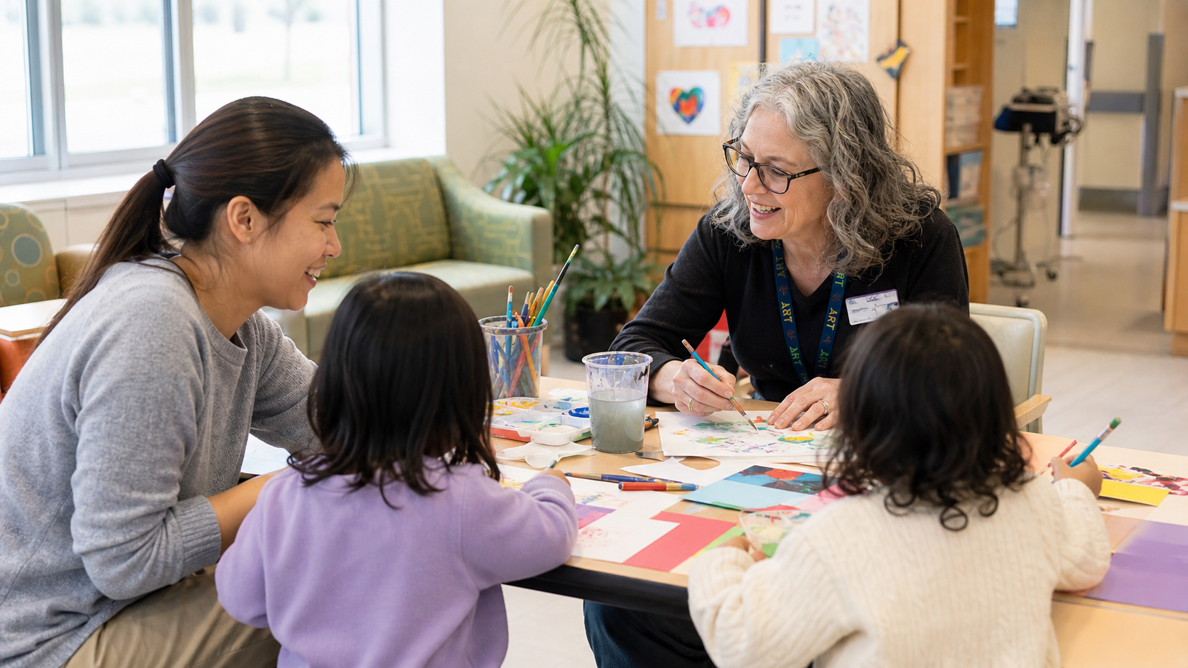 Two adults and two children sitting at a table engaged in a painting activity at a community center or classroom, with art supplies and colorful artwork on the walls.