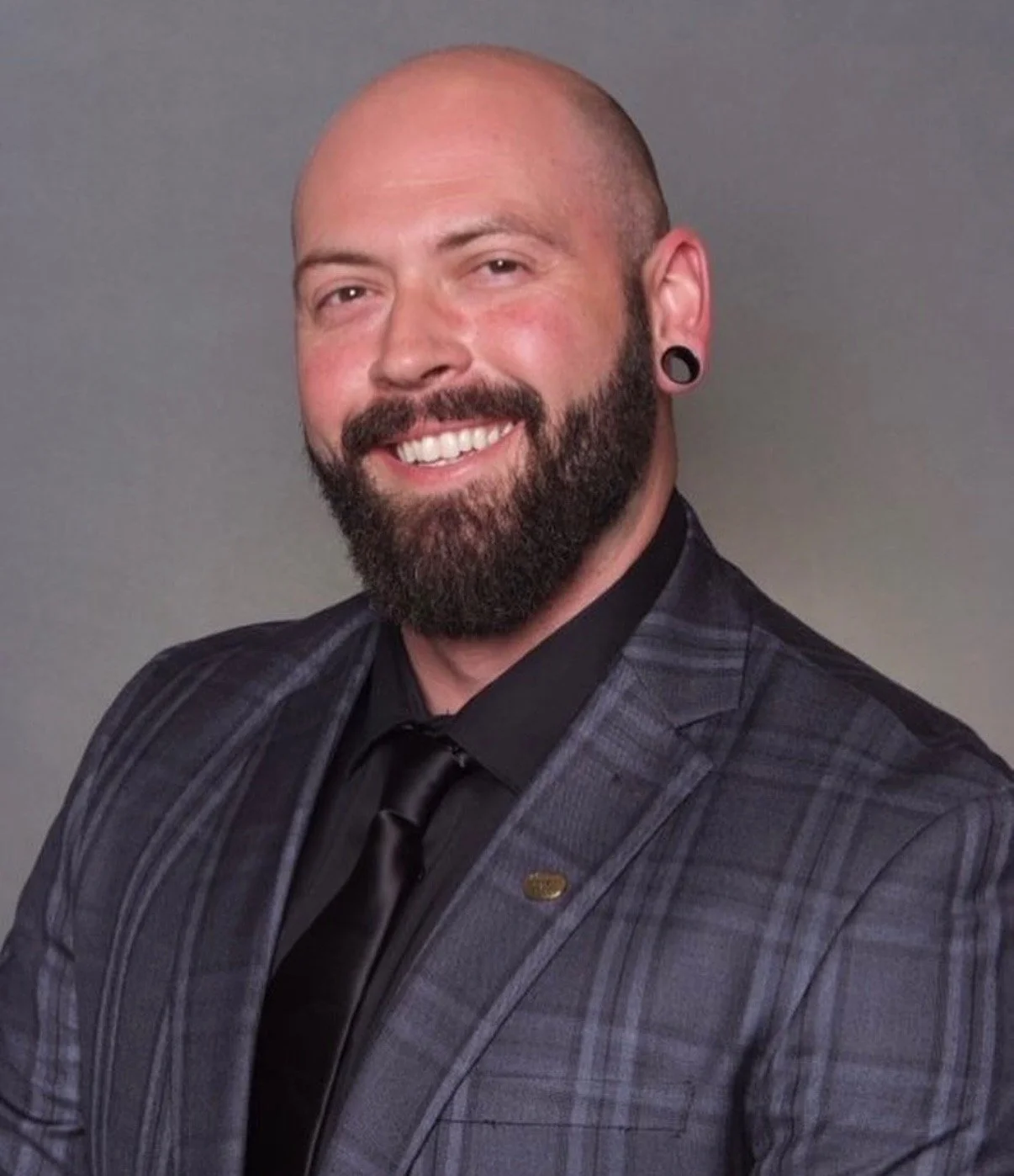 A smiling man with a beard and large black earrings, wearing a plaid suit jacket and black shirt with a black tie, posing against a plain gray background.