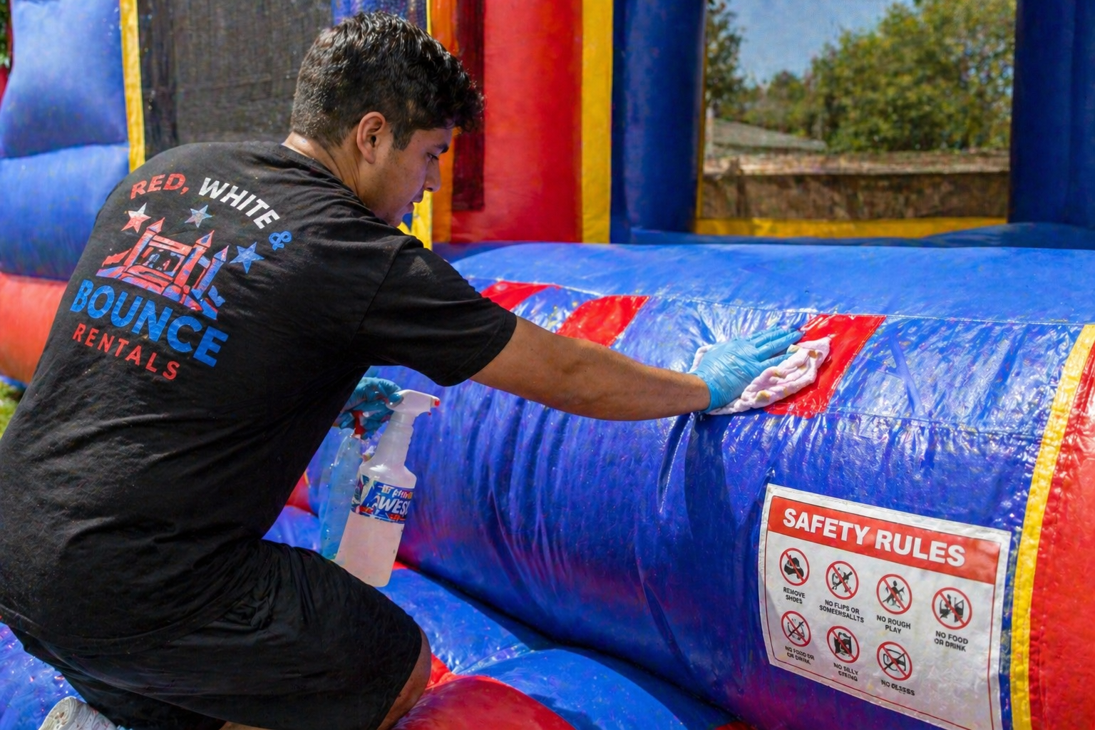 A man cleaning a blue inflatable bounce house with a cloth and spray bottle, wearing a black T-shirt that says 'Red, White & Bounce Rentals'. The bounce house has a safety rules sign on it.