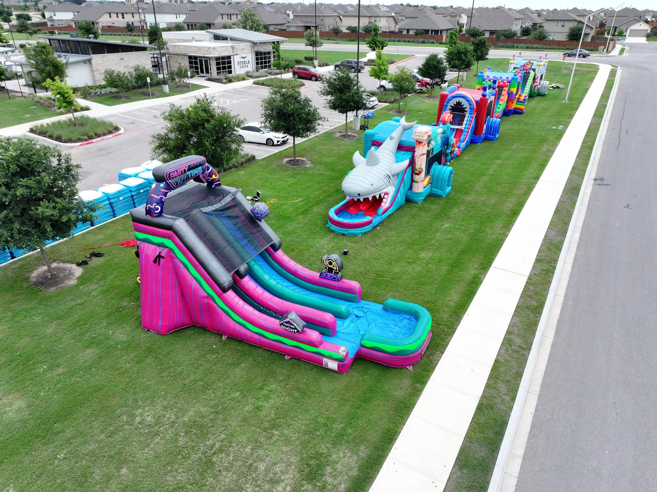 Colorful inflatable water slides and bounce houses set up on a grassy area in a residential neighborhood, with trees, parked cars, and apartment buildings in the background.