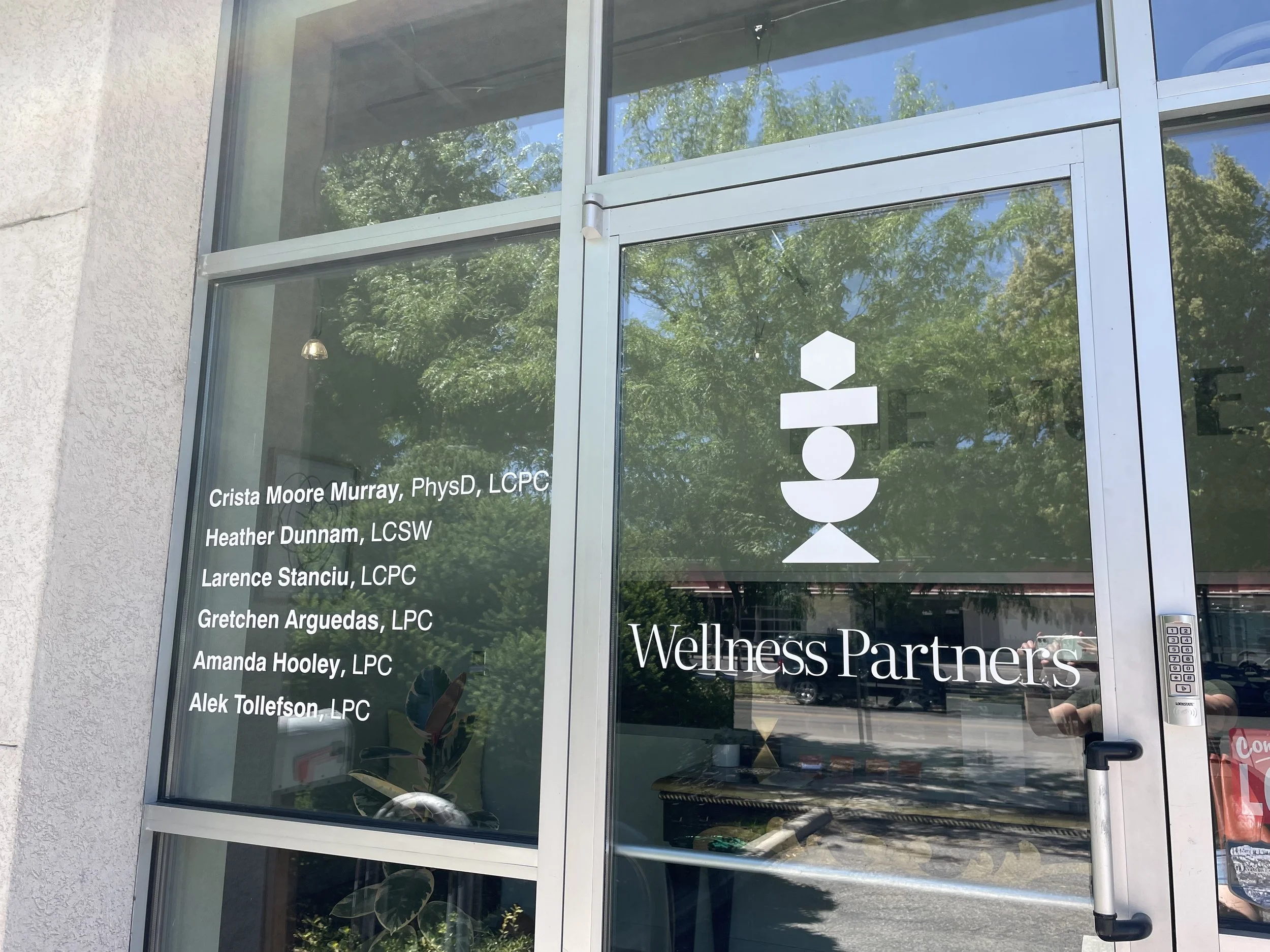 Glass door with a list of names and titles, and the words 'Gretchen Arguedas, LPC'' visible. Reflection of trees and sky in the glass.