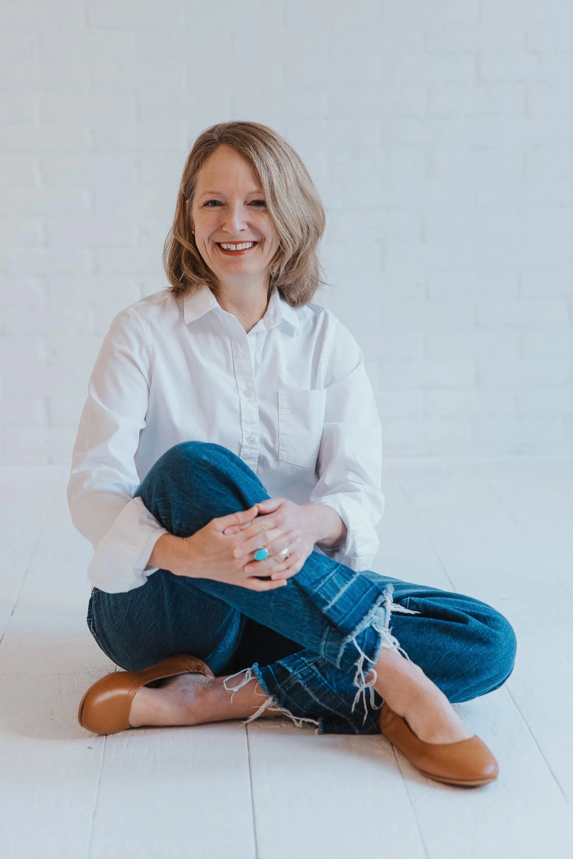 Smiling woman with shoulder-length hair in a white shirt, sitting on the floor with her legs crossed and holding a person in casual jeans and brown shoes. The background is a white brick wall.