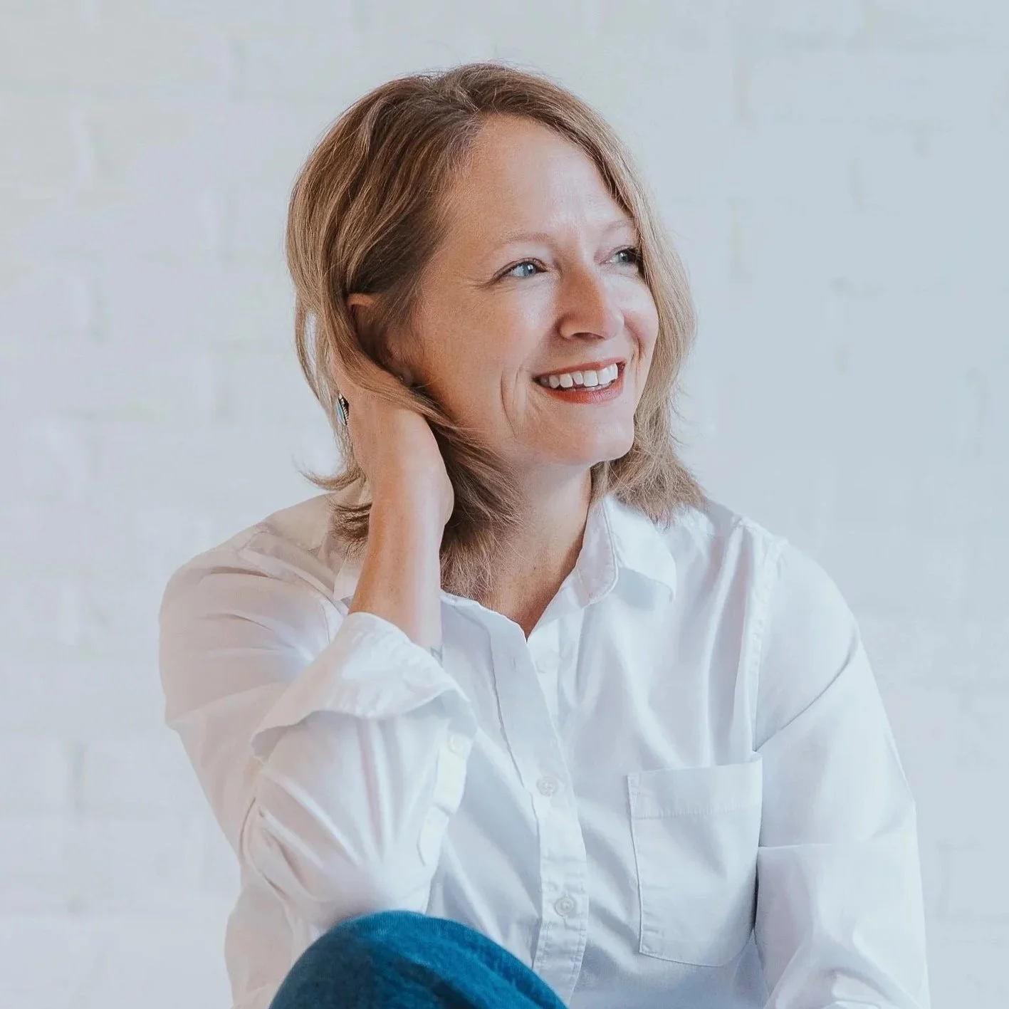 A woman with shoulder-length light brown hair, wearing a white shirt, smiling and looking to her right with her left hand touching her ear, against a light-colored background.