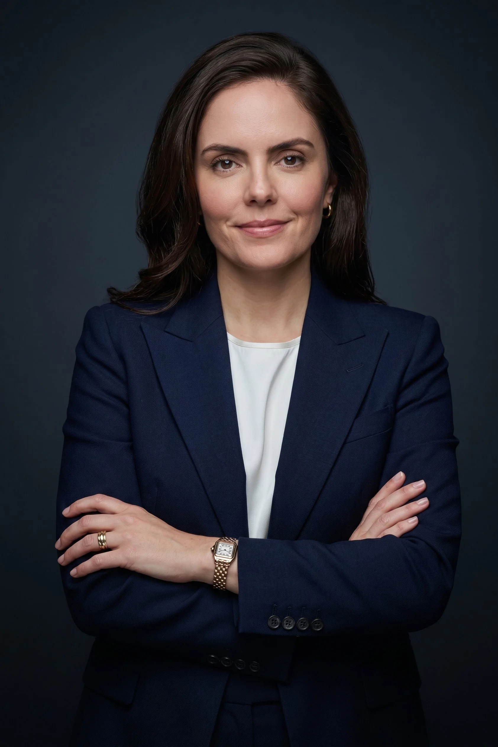 Mulher com cabelo castanho escuro, usando blazer azul escuro e camisa branca, com expressão confiante, braços cruzados, relógio dourado, anel na mão esquerda, fundo escuro.