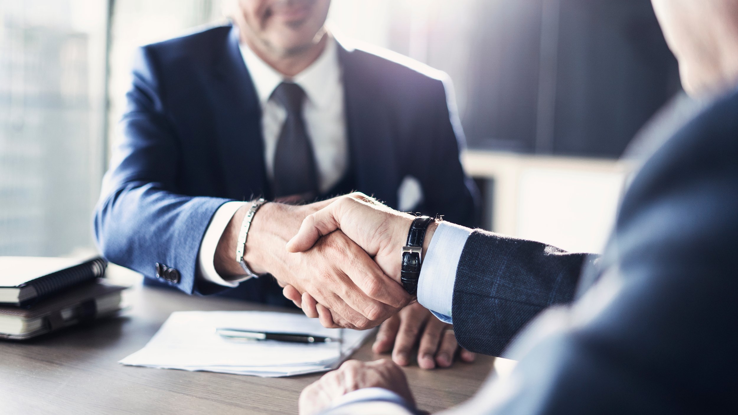 Duas pessoas em roupa formal dando um aperto de mão sobre uma mesa, em ambiente de escritório.