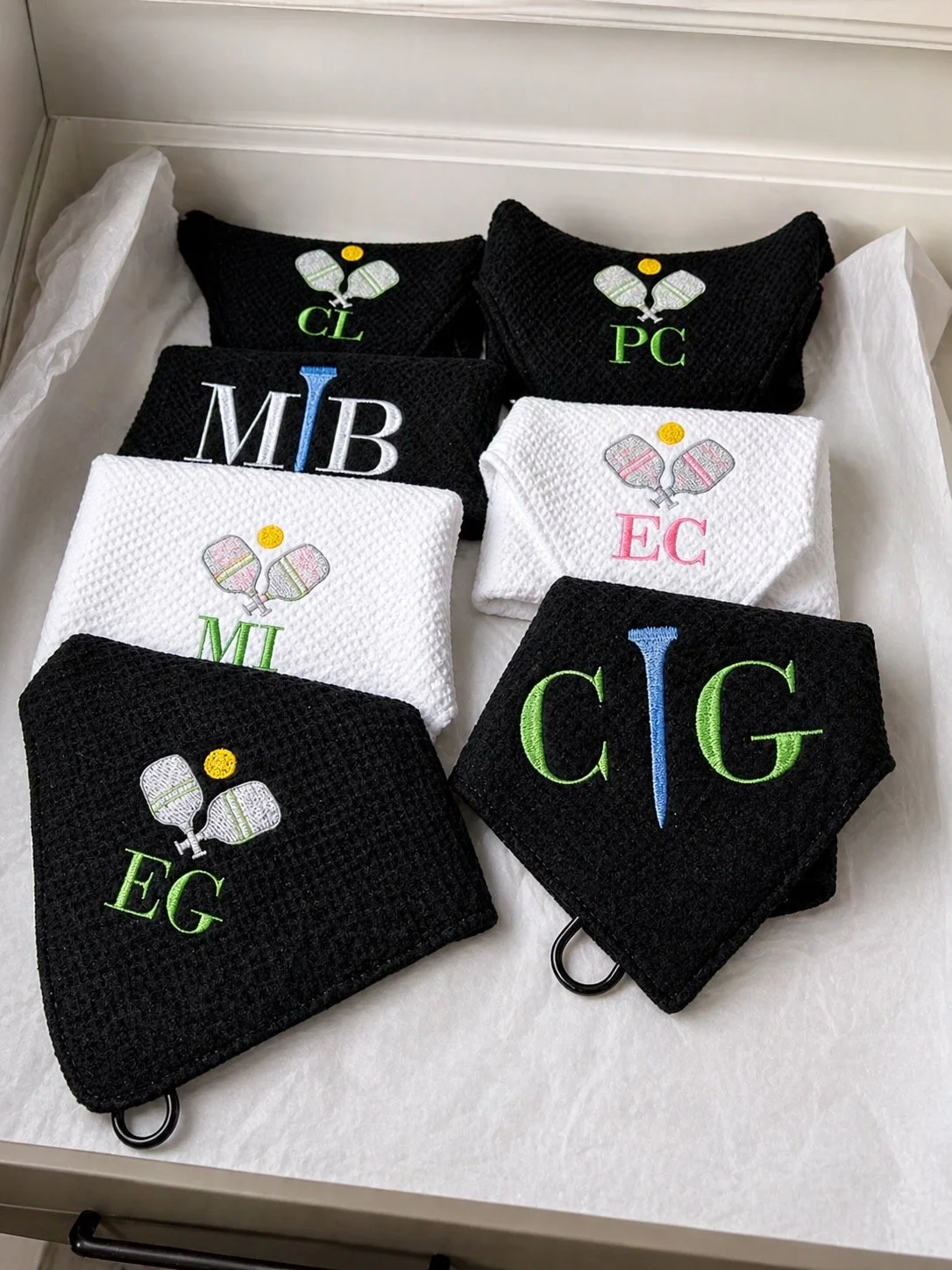 A collection of embroidered towels with various Greek letters and tennis-themed designs, placed inside a box lined with tissue paper.