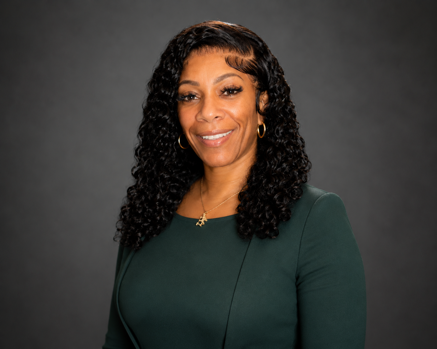 A smiling woman with curly black hair wearing a dark green dress, gold hoop earrings, and a gold necklace with a pendant, posing against a dark gray background.