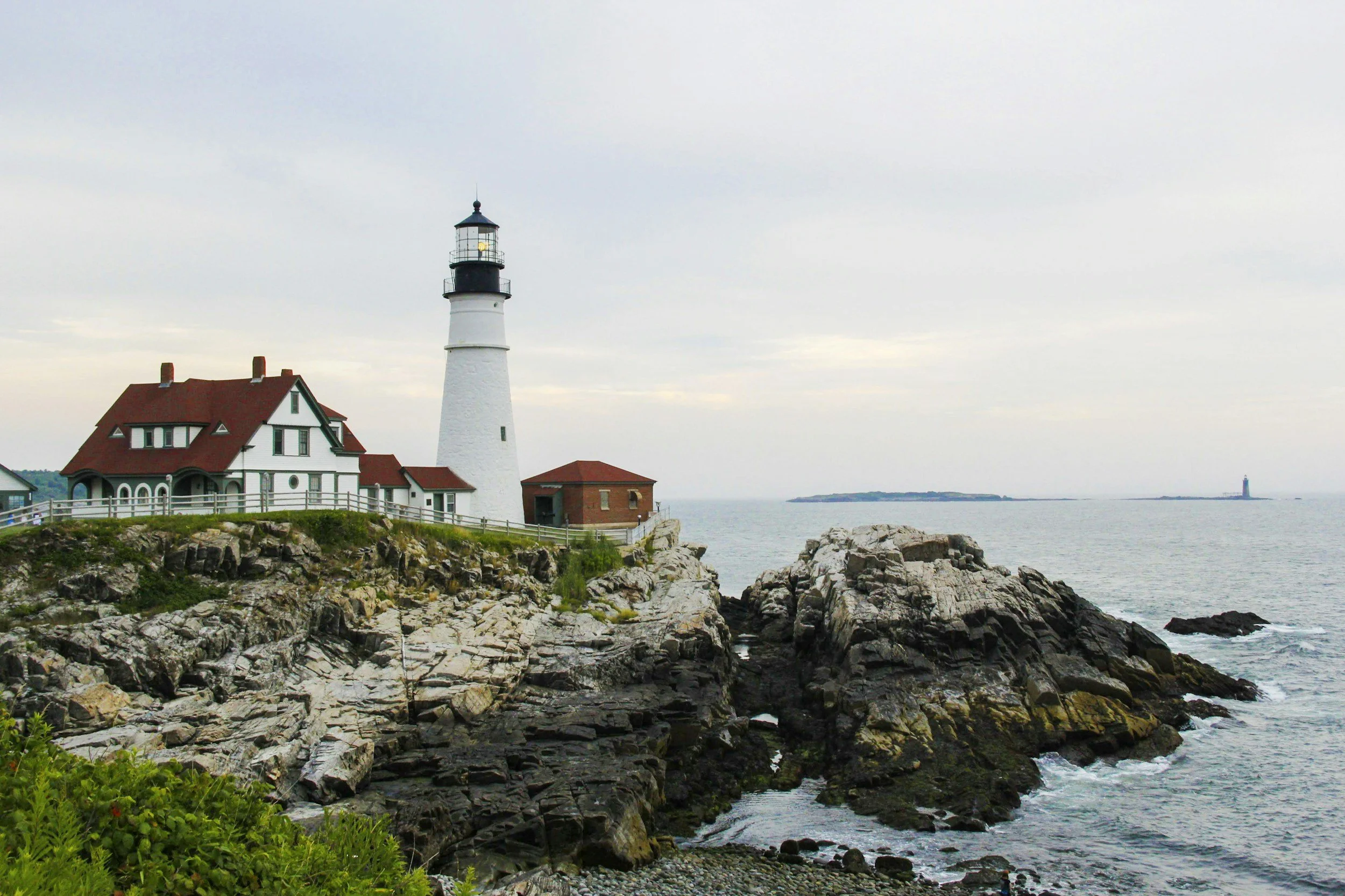 A lighthouse standing next to a house with a red roof on a rocky shoreline, with water and another lighthouse in the distance under a cloudy sky.