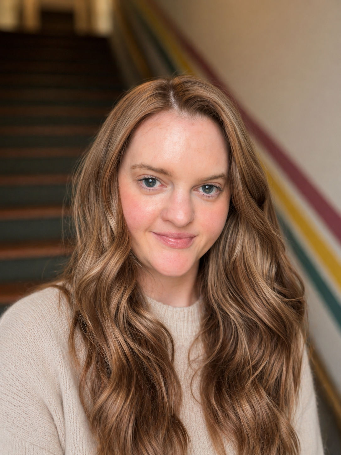 A woman with long wavy light brown hair and blue eyes smiling at the camera, standing indoors near colorful stairs.