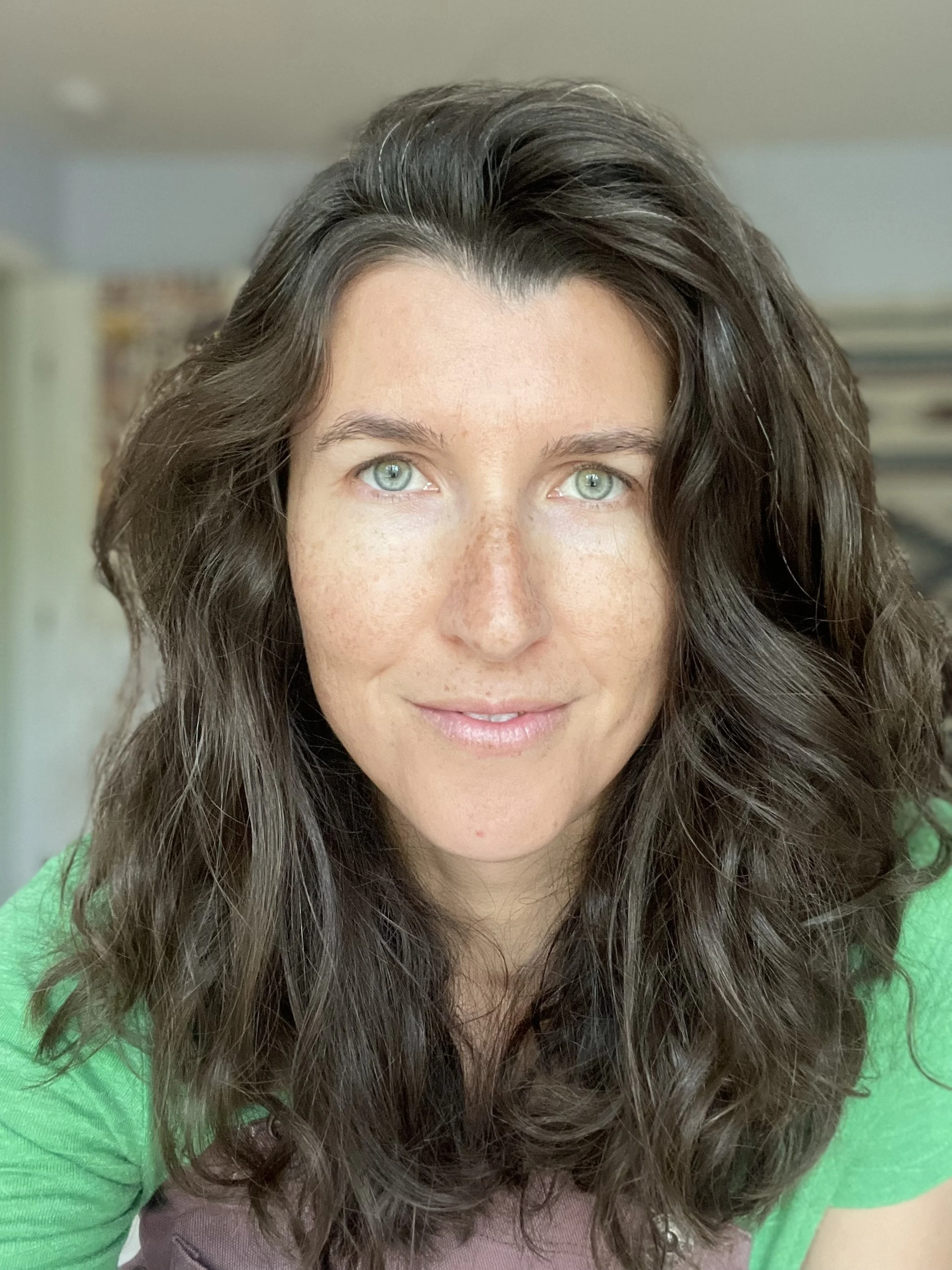 Close-up of a woman with curly brown hair, fair skin, and blue eyes, wearing a green top, smiling gently.