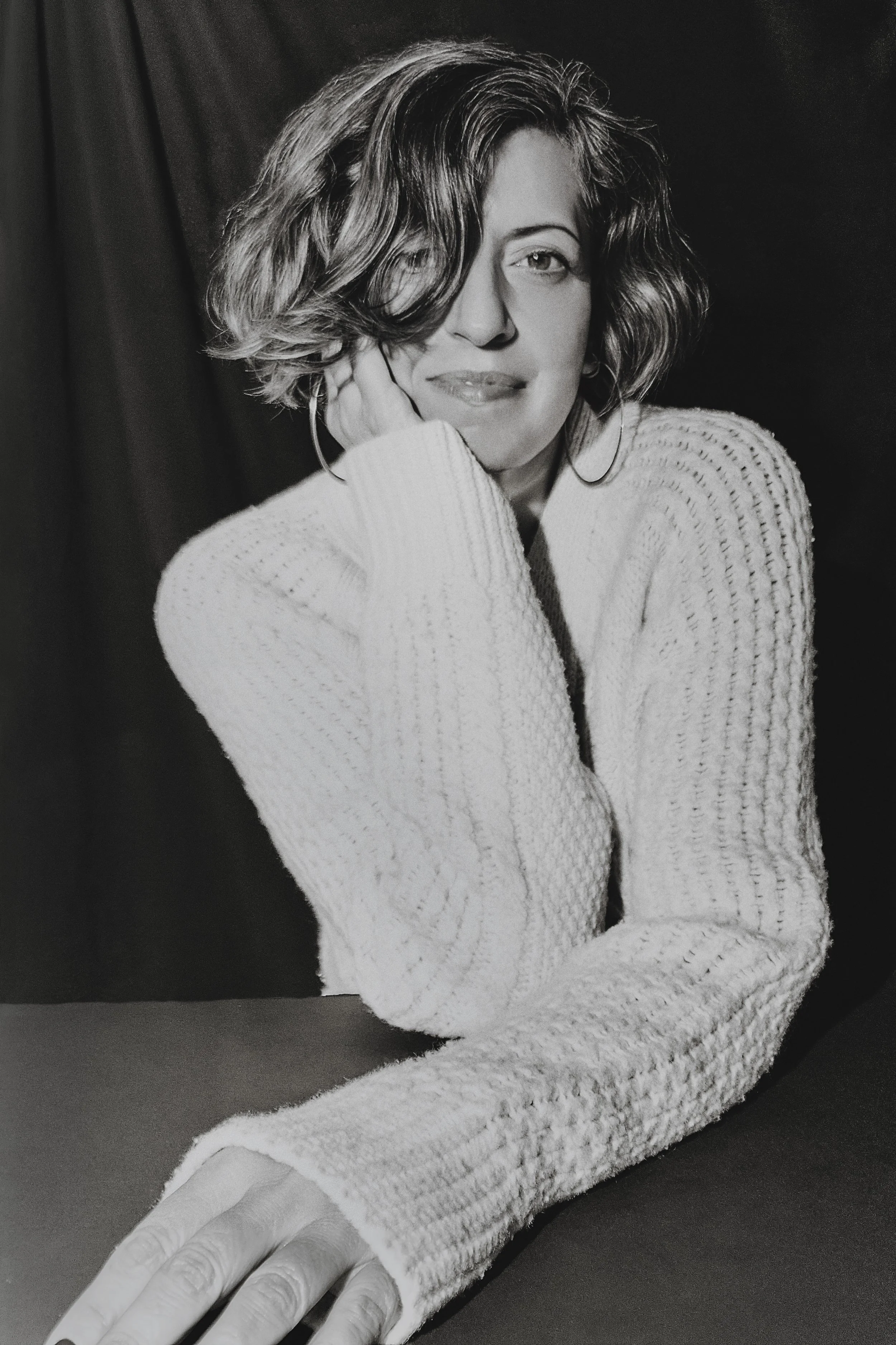 Portrait of a woman with wavy hair, wearing a knit sweater, resting her hand on her cheek, against a dark background.