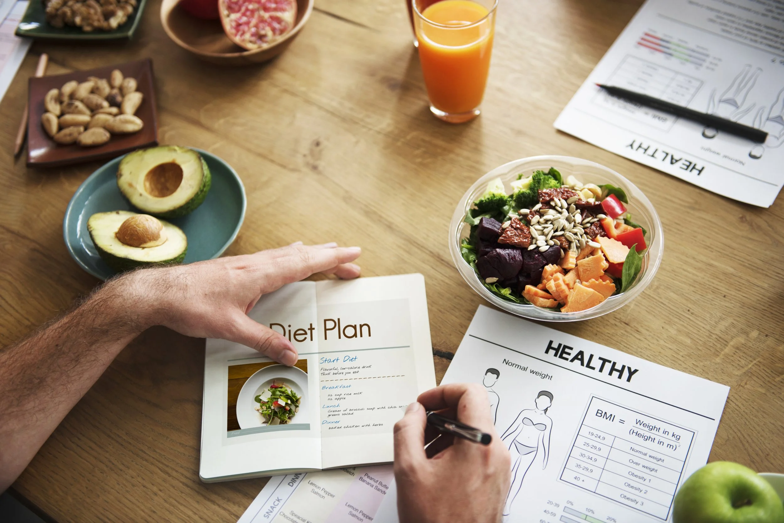A person holding a dietary plan sheet with a salad, an avocado, and various healthy food items on a wooden table, accompanied by food and drink items, including a juice glass and nuts.