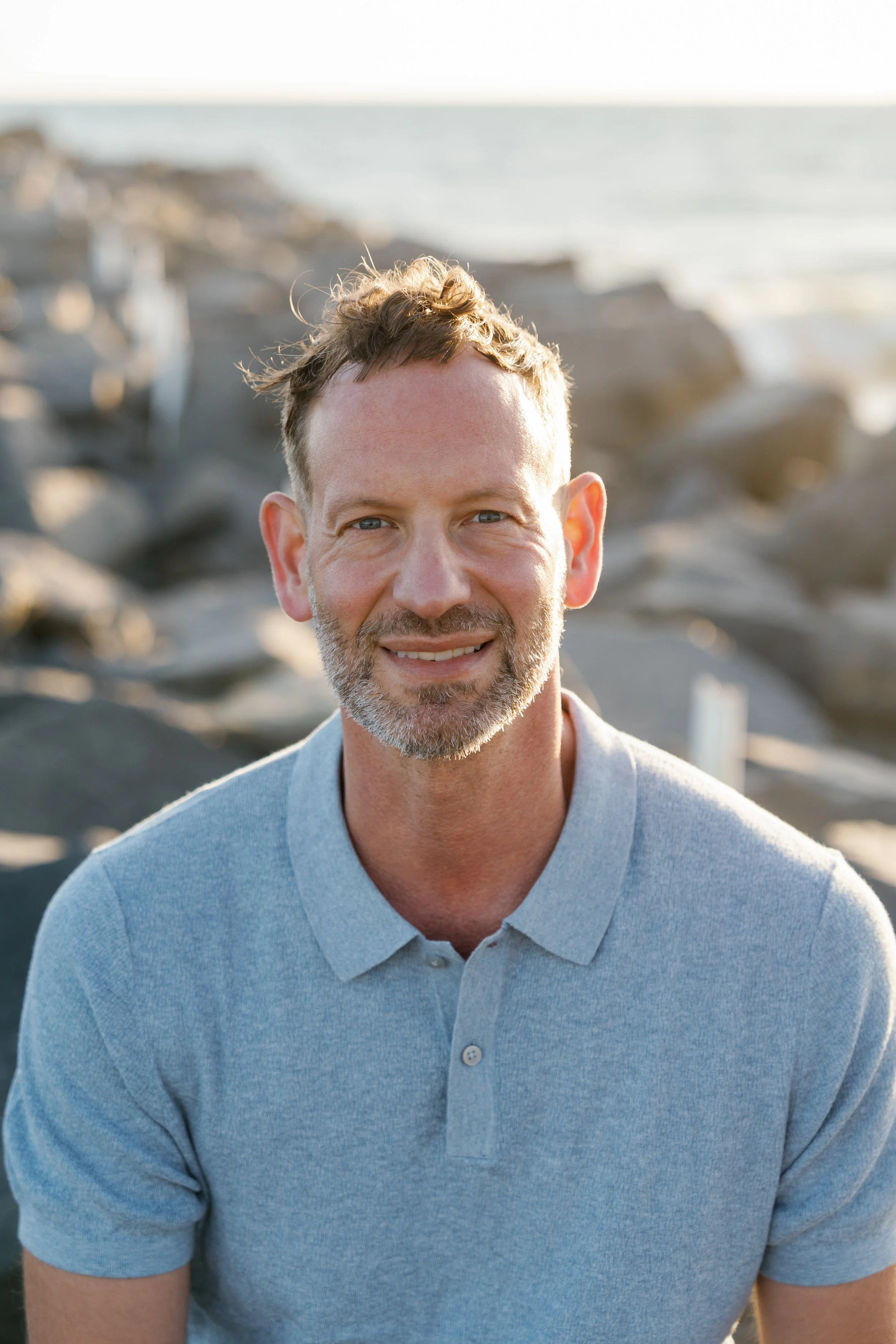 A man with short, light brown hair and a beard, wearing a light blue polo shirt, smiling at the camera on a rocky beach during sunset.