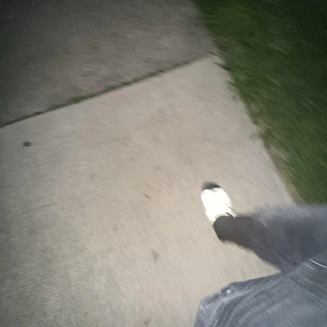 A person walking on a sidewalk at night, wearing black pants and a white sneaker, with grass on the side.