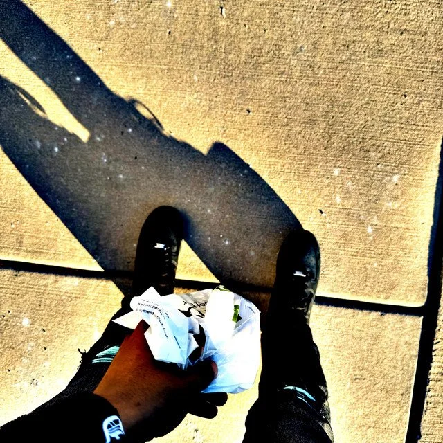 Photo taken from above showing a person wearing black shoes, black socks, and black pants holding crumpled paper bag on a sidewalk with their shadow in the sunlight.