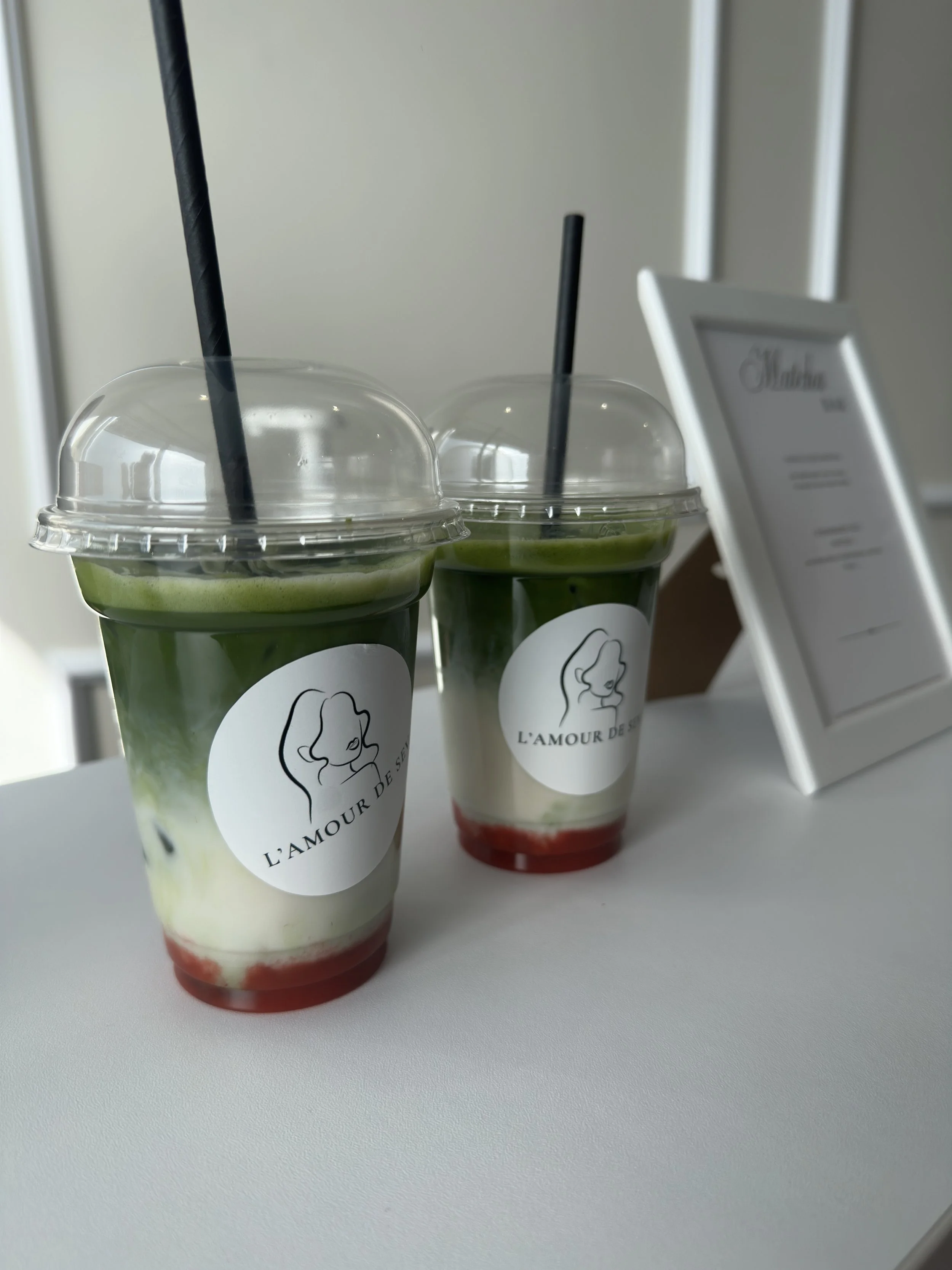 Zwei grüne Smoothies in transparent Bechern mit schwarzen Strohhalmen und einem weißen Logo, daneben ein kleines Whiteboard mit dem Schriftzug 'L'AMOUR DE S'.