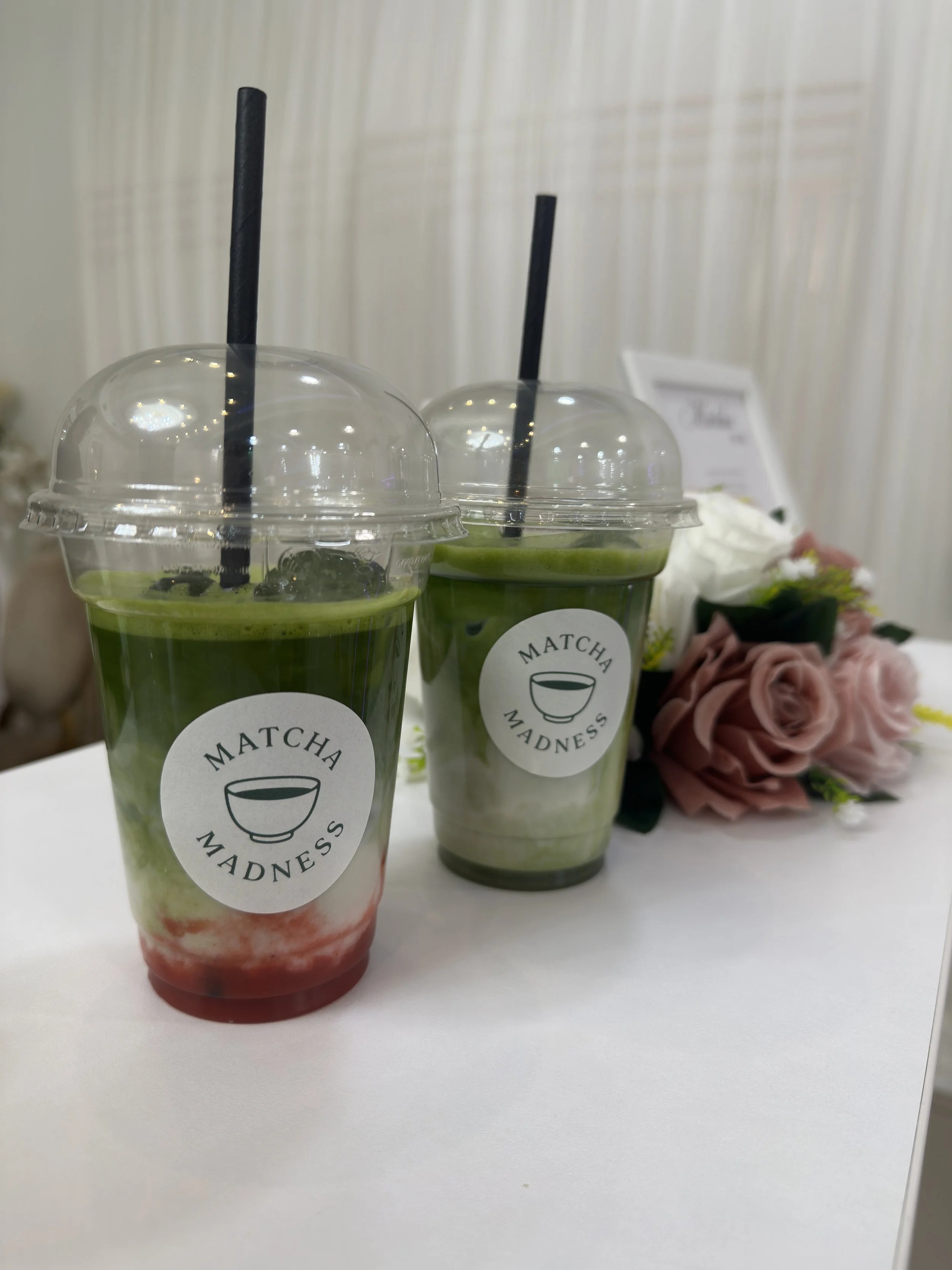 Zwei grüne Matcha-Getränke mit schwarzen Strohhalmen, auf einem weißen Tisch, im Hintergrund eine Blumenarrangement.