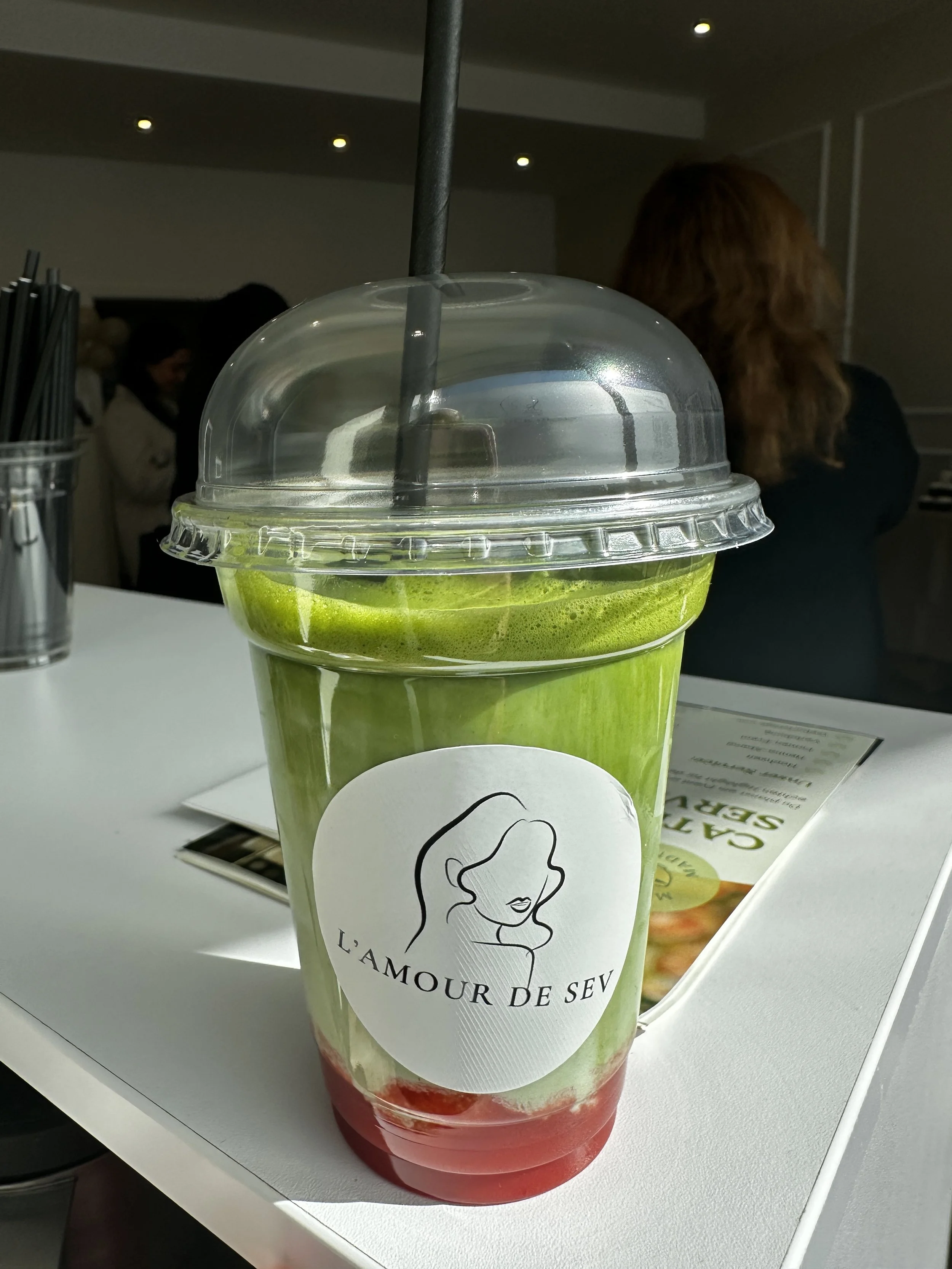 Ein grün-roter Fruchtcocktail in einem transparenten Becher mit einem schwarzen Strohhalm, an einem weißen Tisch im Café L'Amour de Seveu.