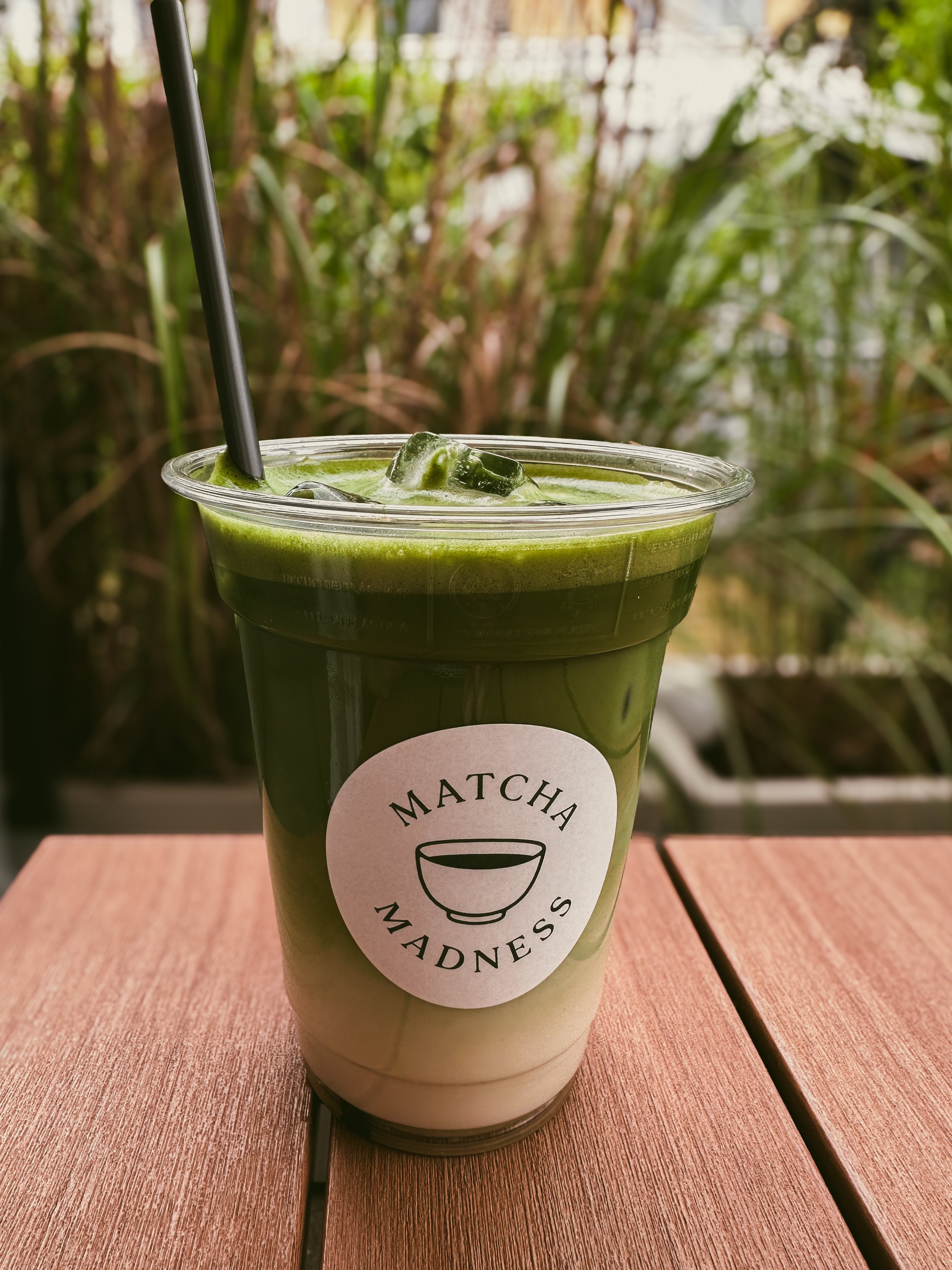 Ein grünes Matcha-Getränk in einem transparenten Becher mit Eisklumpen und einem schwarzen Strohhalm, auf Holz Tisch mit grüner Pflanzenhintergrund.
