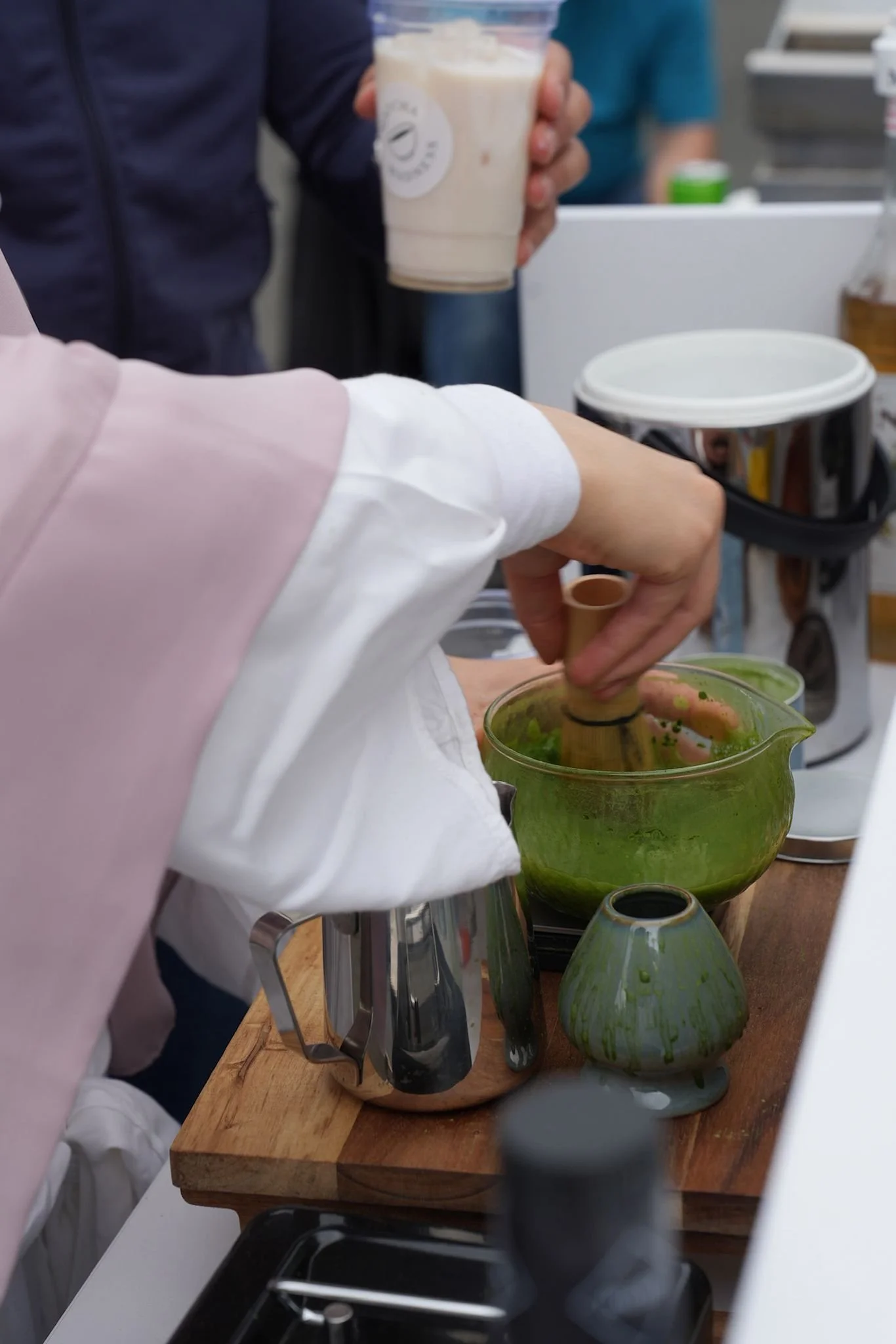Person, die ein grünes Pesto in einem Glas rührt, während jemand anderes ein Getränk hält, vermutlich in einer Küchenszene.