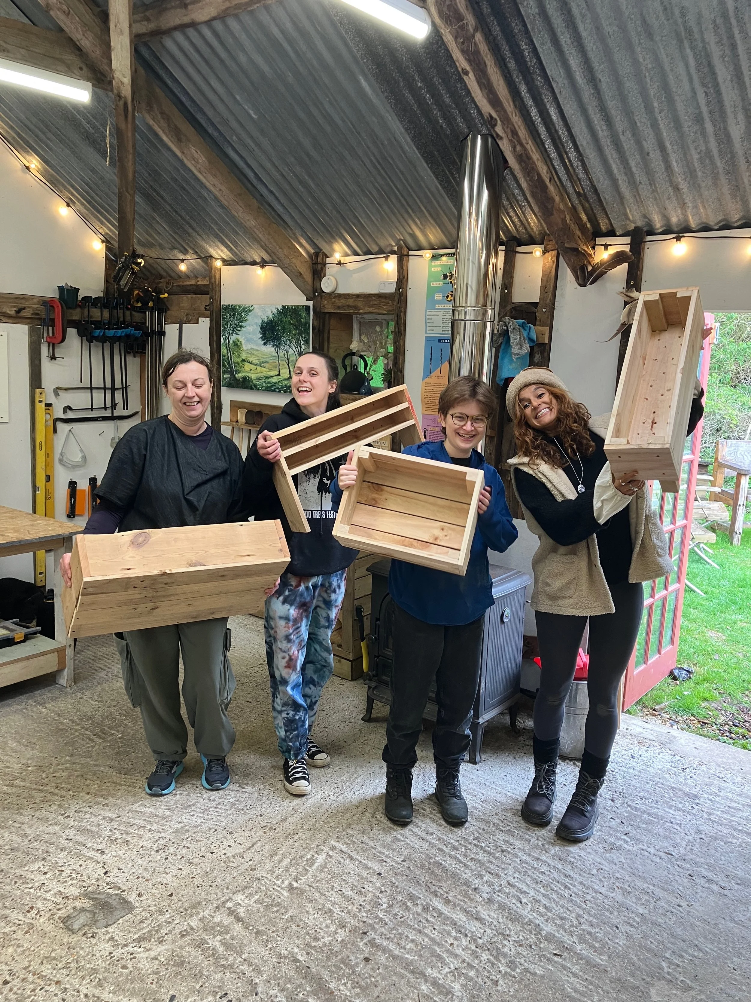 Four women and non-binary people smiling and holding wooden crates inside a workshop with tools on the wall and a painting of trees in the background.