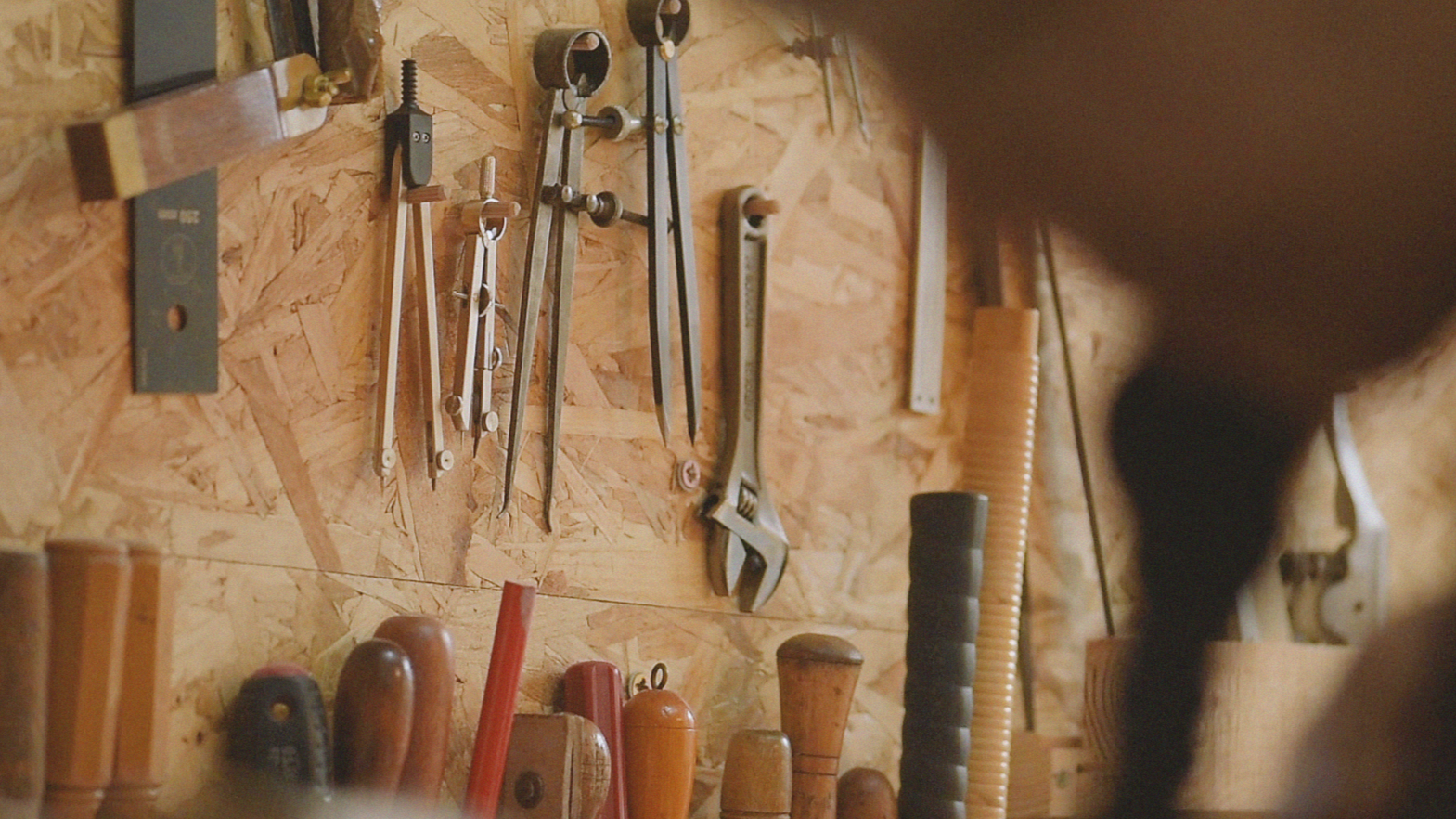 A woodworking workshop wall with various hand tools hanging, including compasses, a wrench, and a level. Below, there are wooden handles and tools placed on a workbench.