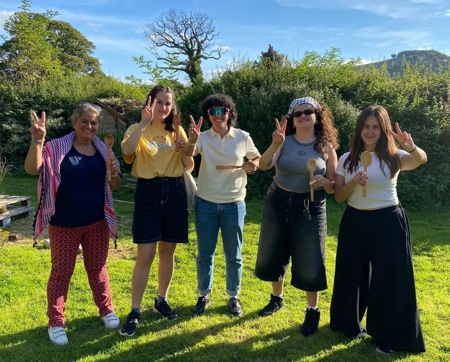 A group of five women standing outdoors on grass, smiling, holding wooden spoons, and making peace signs with their fingers, with trees and a blue sky in the background.