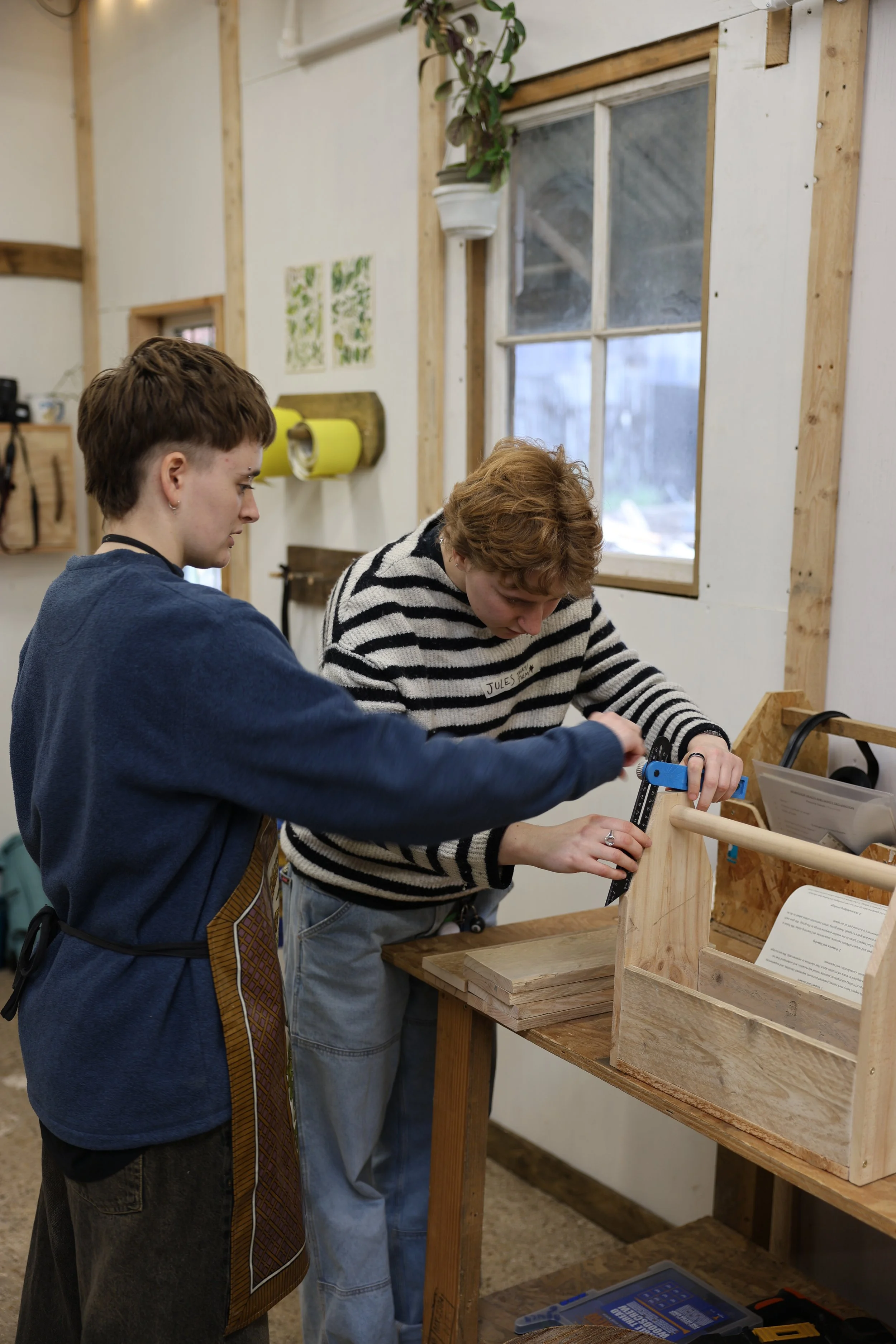 Two people working on a woodworking project at a workbench inside a workshop. One is holding a tool, and the other is assisting. The workshop has a window, a potted plant, and various tools and materials on the walls.