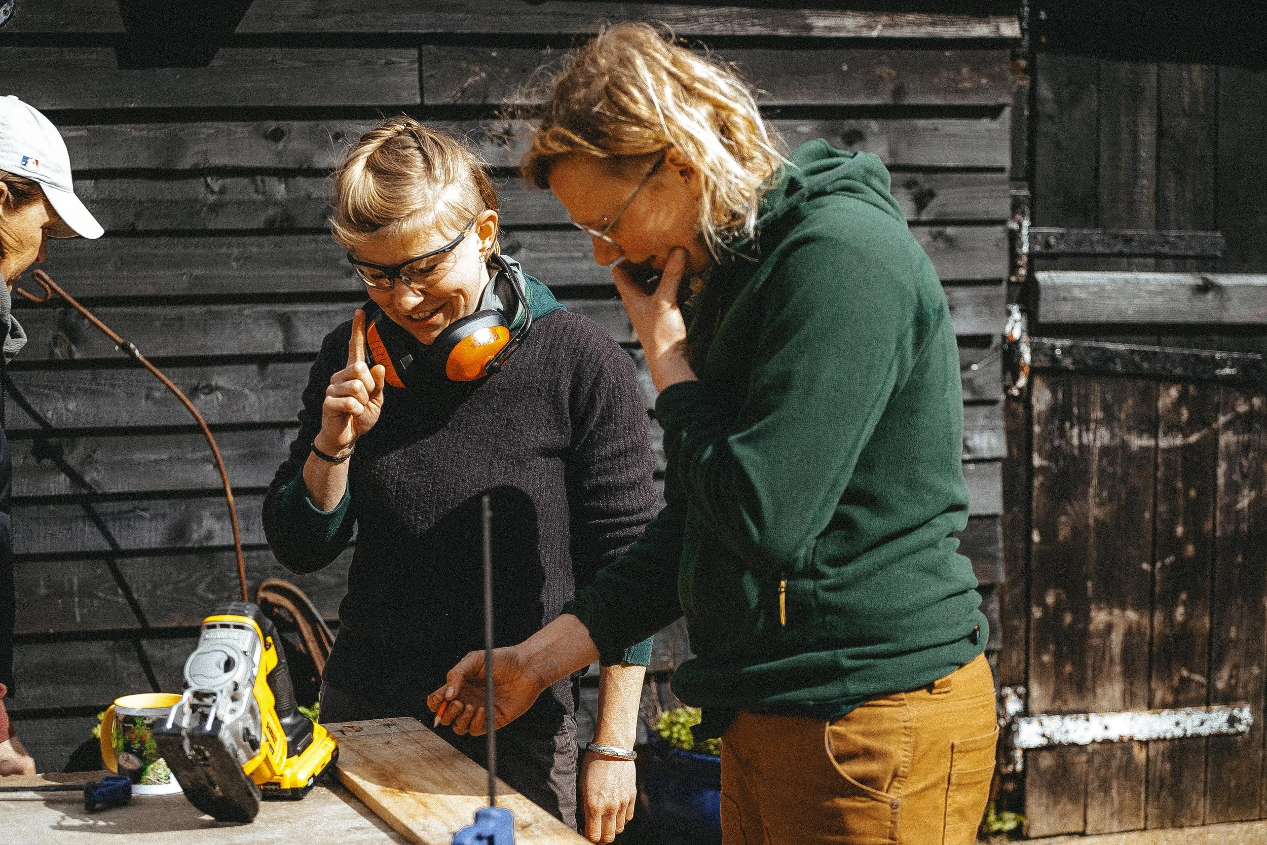 Three people are gathered around a wooden workbench outside, with a black wooden fence in the background. One person is wearing a white cap, another has glasses and a headset around her neck, and the third has wavy blonde hair, glasses, and is wearing a green hoodie. They appear to be discussing or working on something together, with tools and equipment on the table, including a yellow power tool.