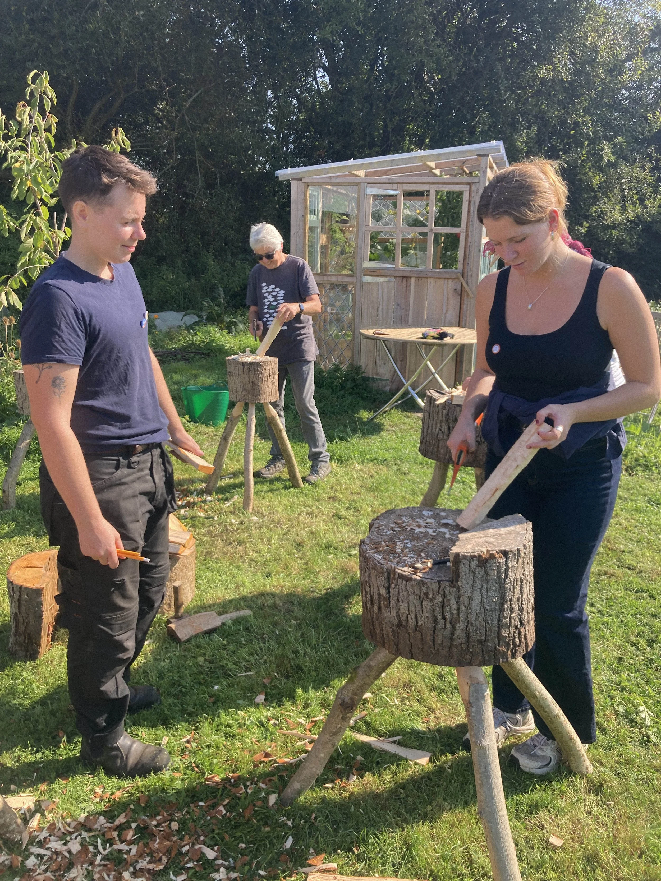 Three people outdoors chopping wood, with a wood shed in the background and green trees surrounding them.