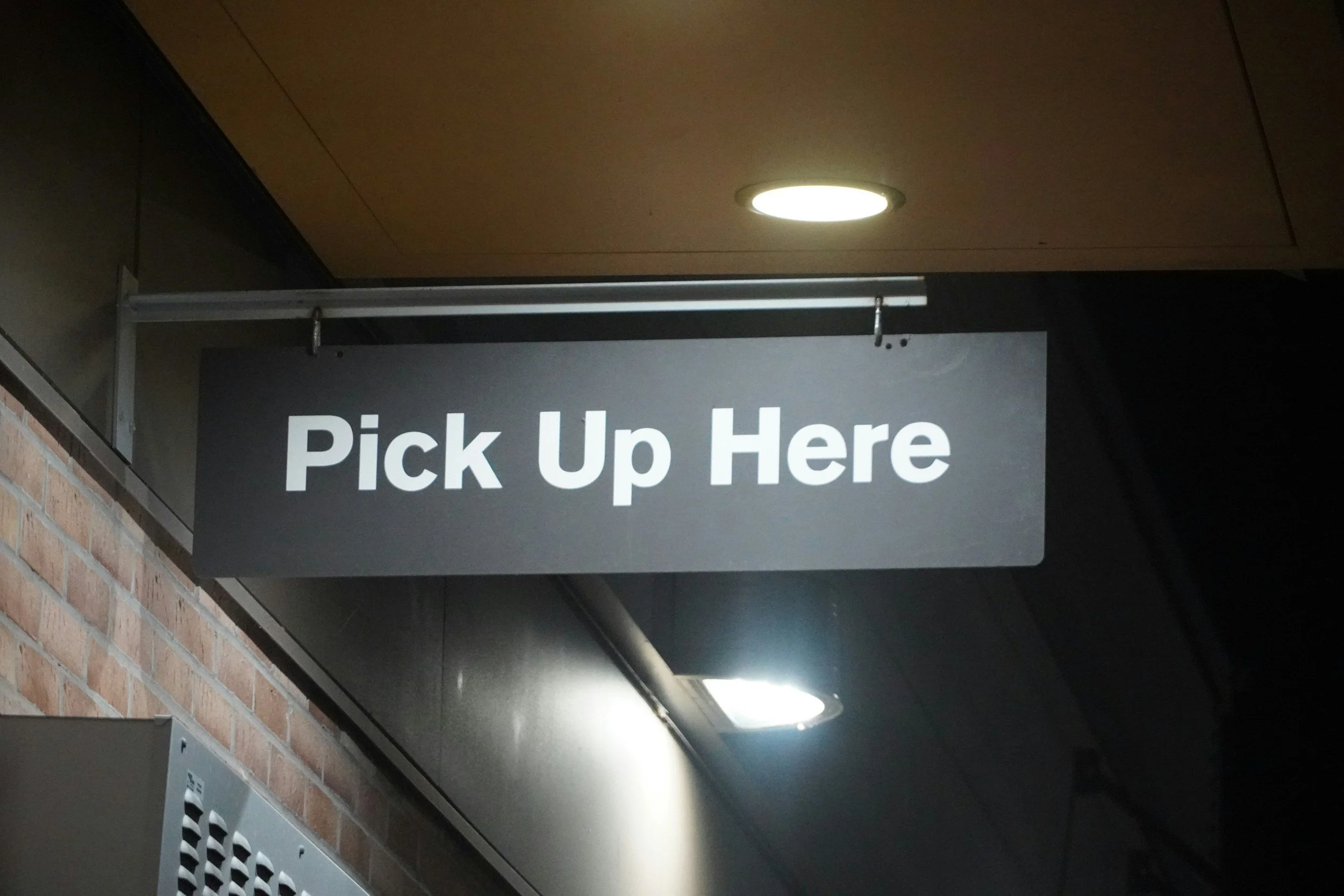 Black sign with white text that reads 'Pick Up Here' hanging from the ceiling in a building corridor.
