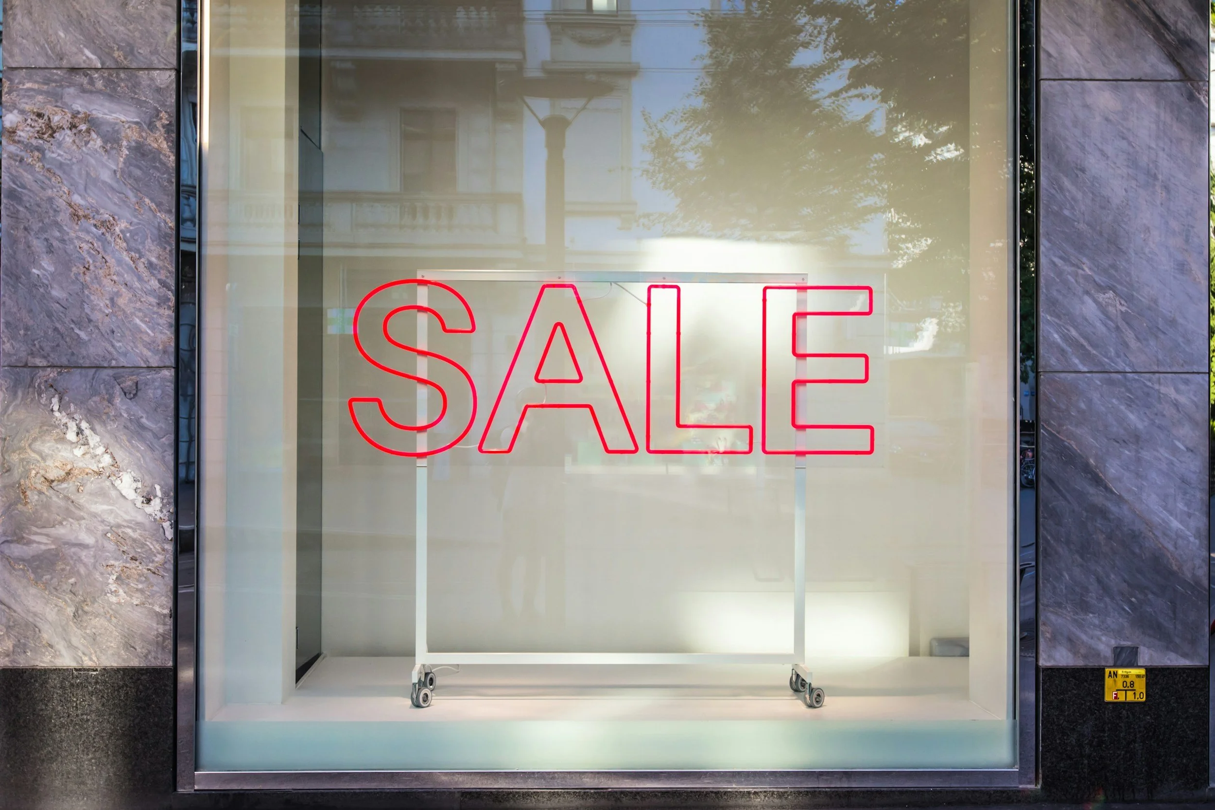 A storefront window with a large red neon sign that reads 'SALE'. The display window is surrounded by a marble-like frame and is empty except for the sign, with a reflection of buildings and trees visible in the glass.