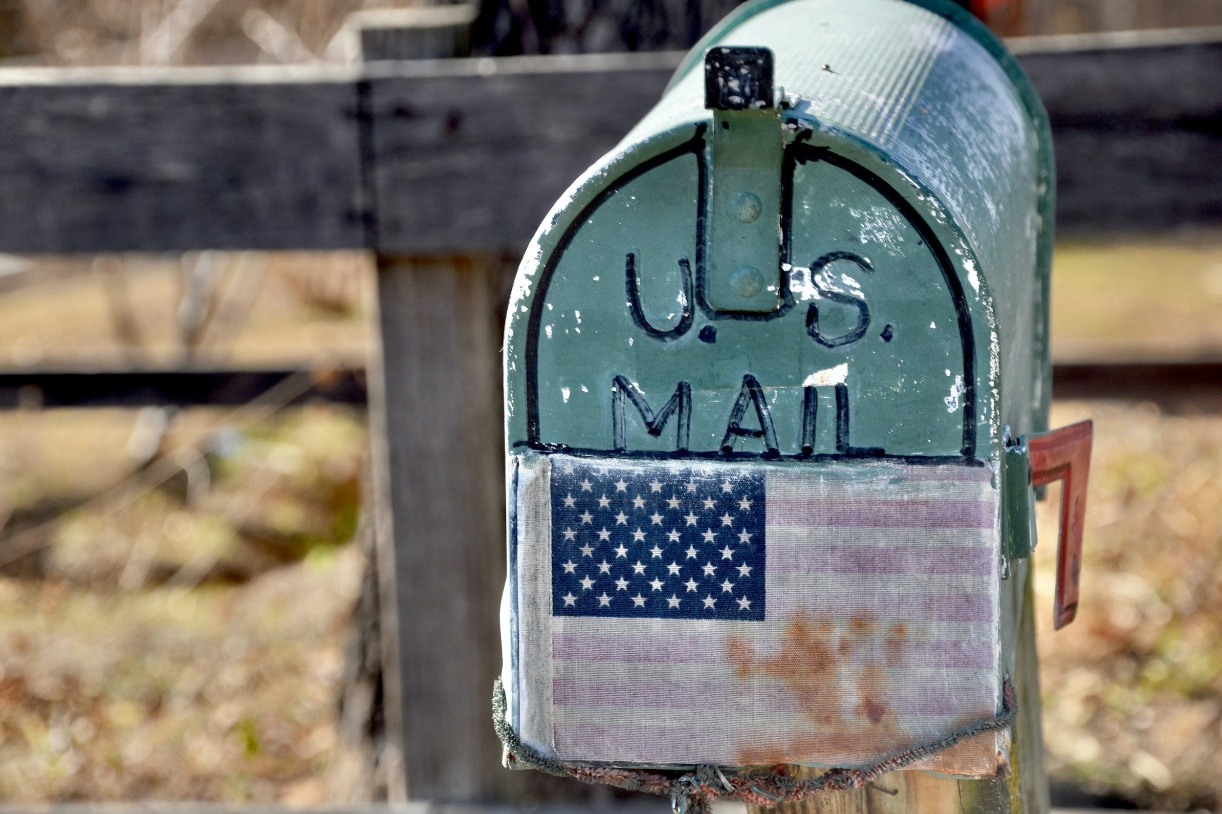 A green mailbox attached to a wooden post with an American flag sticker. The mailbox is weathered and has white lettering that reads 'U.S. MAIL'.