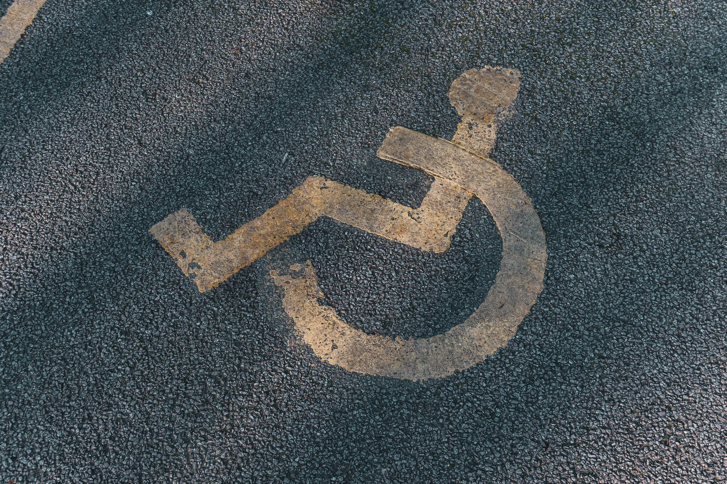 A yellow, handicap symbol painted on a rough asphalt road, with the yellow paint faded and chipped, as if frequently used.