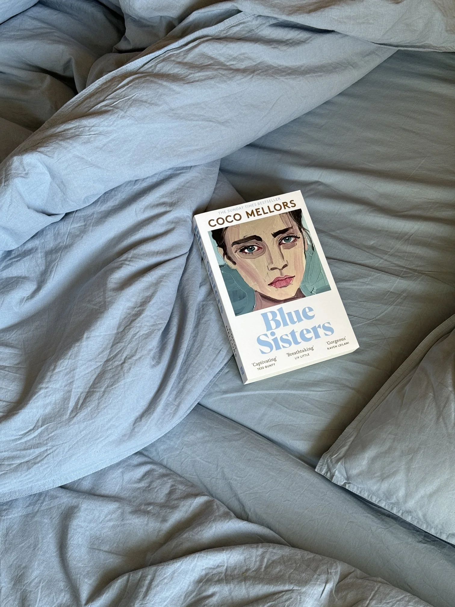 A book titled "Blue Sisters" resting on crumpled light gray bed sheets.