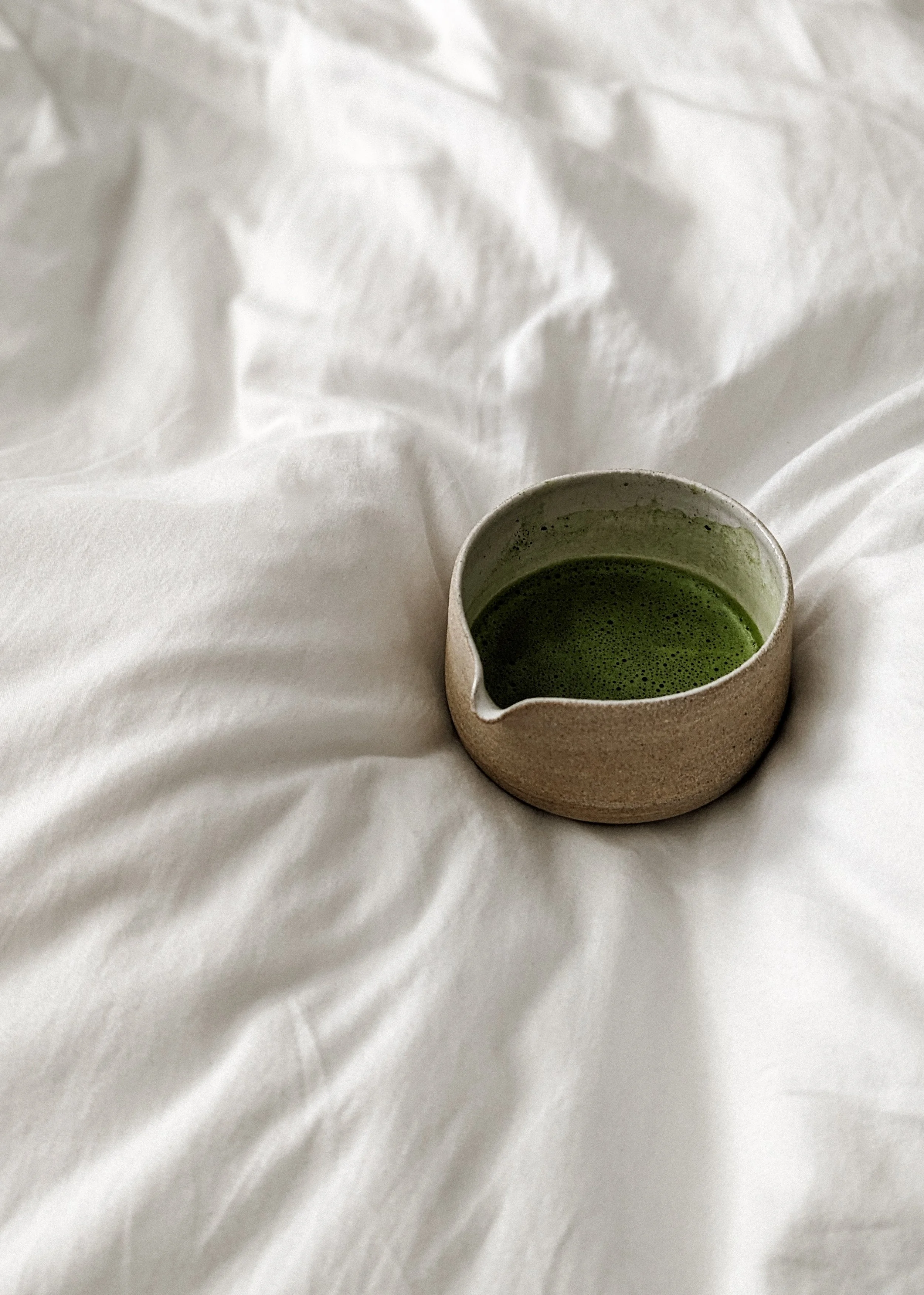 A ceramic cup filled with green matcha tea on a crumpled white bedsheet.