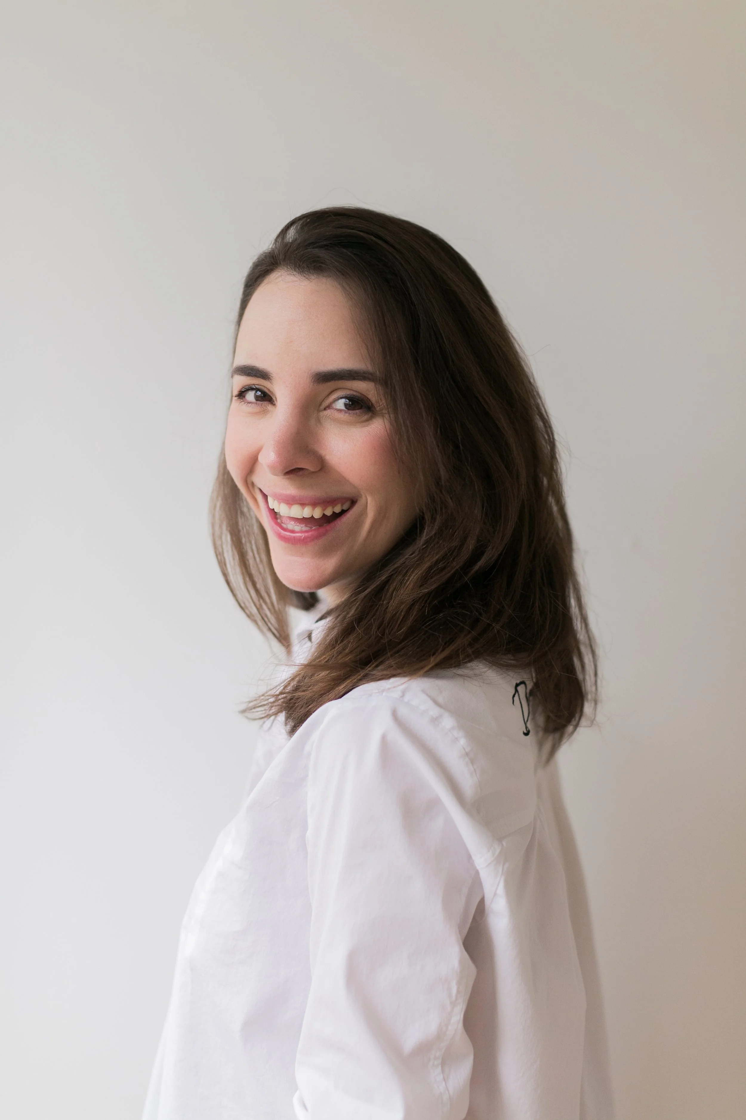 A young woman with brown hair and a big smile, wearing a white shirt, looking at the camera against a plain white background.