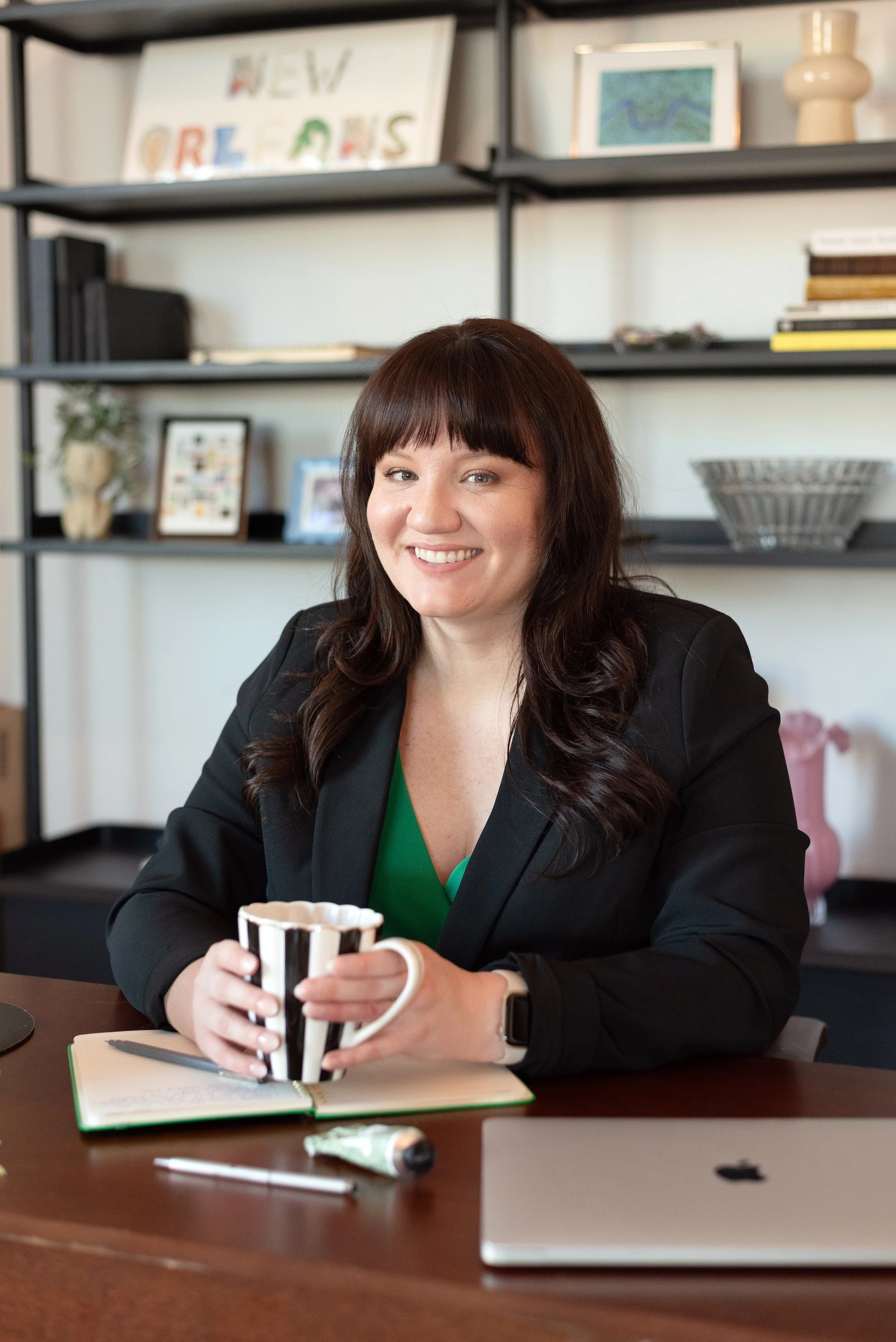 Amelia sits at a desk wearing a black blazer over a green dress.
