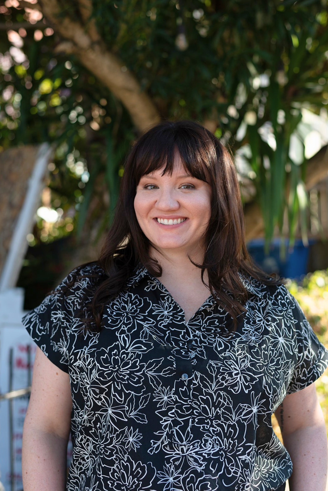 Amelia stands under a tree, smiling at the camera, wearing a black and white printed button up shirt.