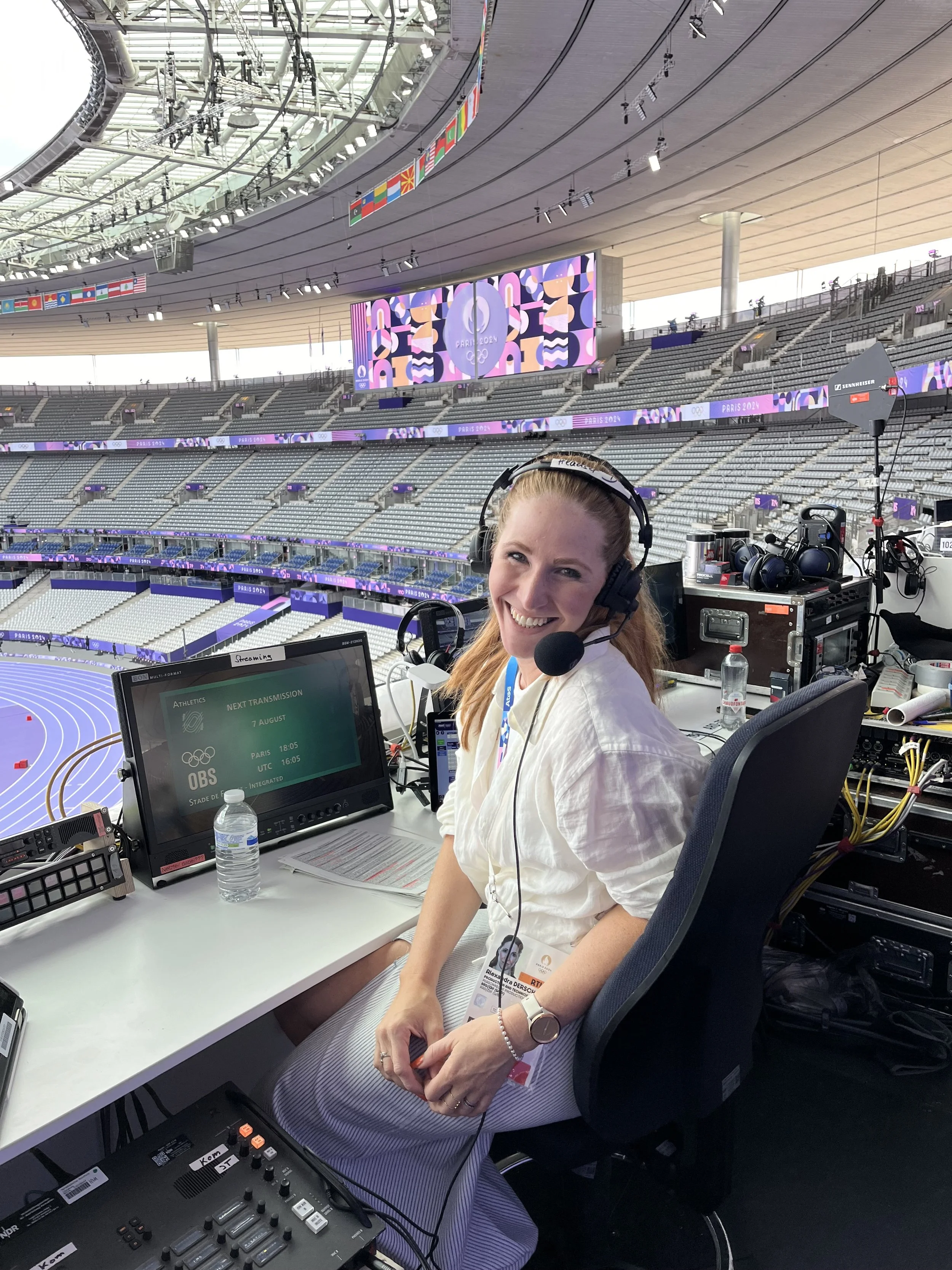 Alexandra Dersch, Moderatorin, Kommentatorin, ZDF, Leichtathletik, Curling, Para Ski Alpin, Skispringen, Sportstudio, Eine Frau sitzt an einem Schreibtisch, trägt Kopfhörer mit Mikrofon, hat ein Lanyard um den Hals und lächelt. Olympia