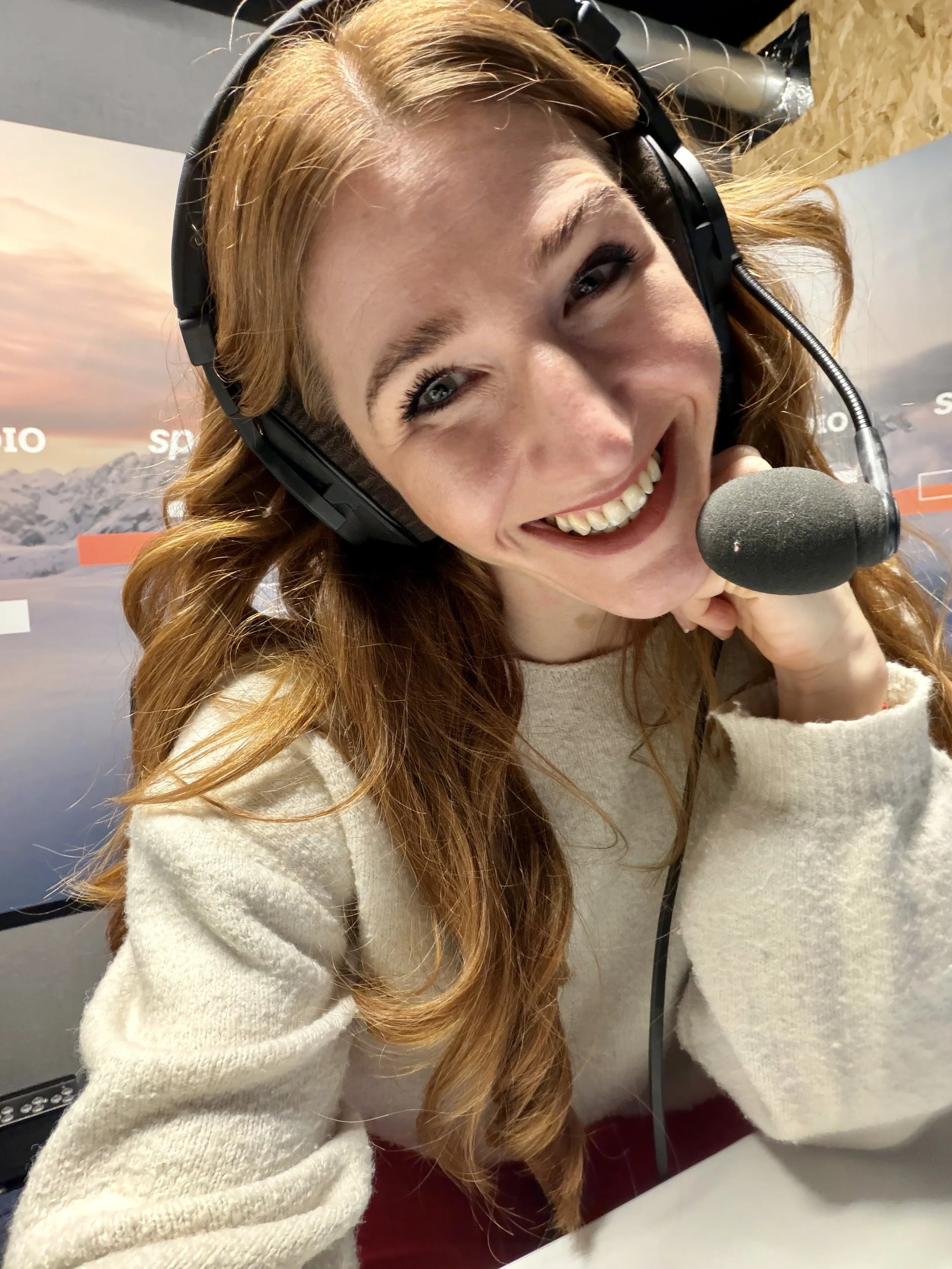 Alexandra Dersch, Moderatorin, Kommentatorin, ZDF, Leichtathletik, Curling, Para Ski Alpin, Skispringen, Sportstudio, Lächelnde Frau mit roten Haaren trägt ein Headset und sitzt an einem Tisch in einem Raum mit Berg-Poster im Hintergrund.