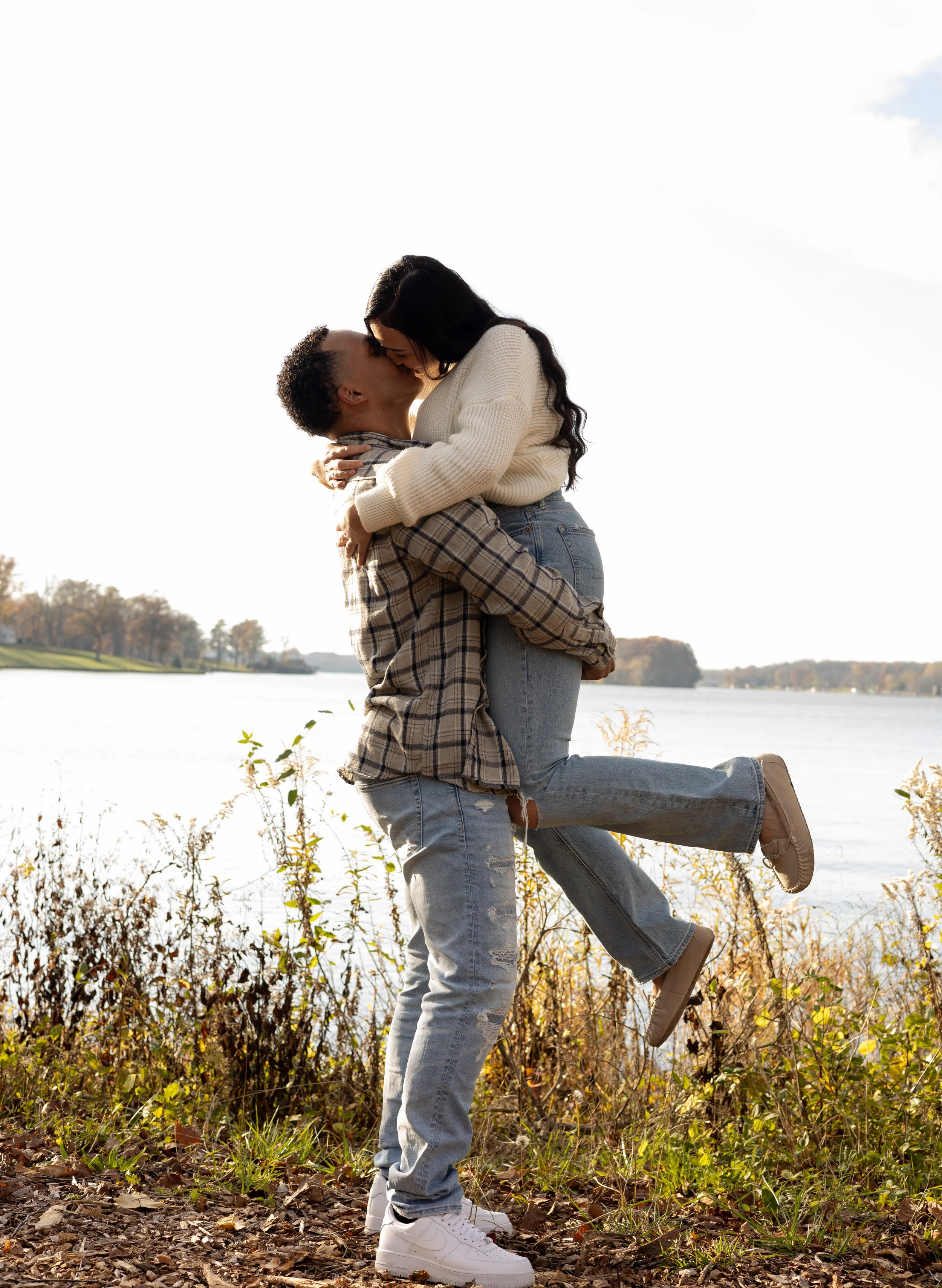A couple standing outdoors, with the man lifting and kissing the woman. They are near a body of water with trees in the background, and the woman is wearing a white sweater and jeans, while the man is wearing a plaid shirt and jeans.