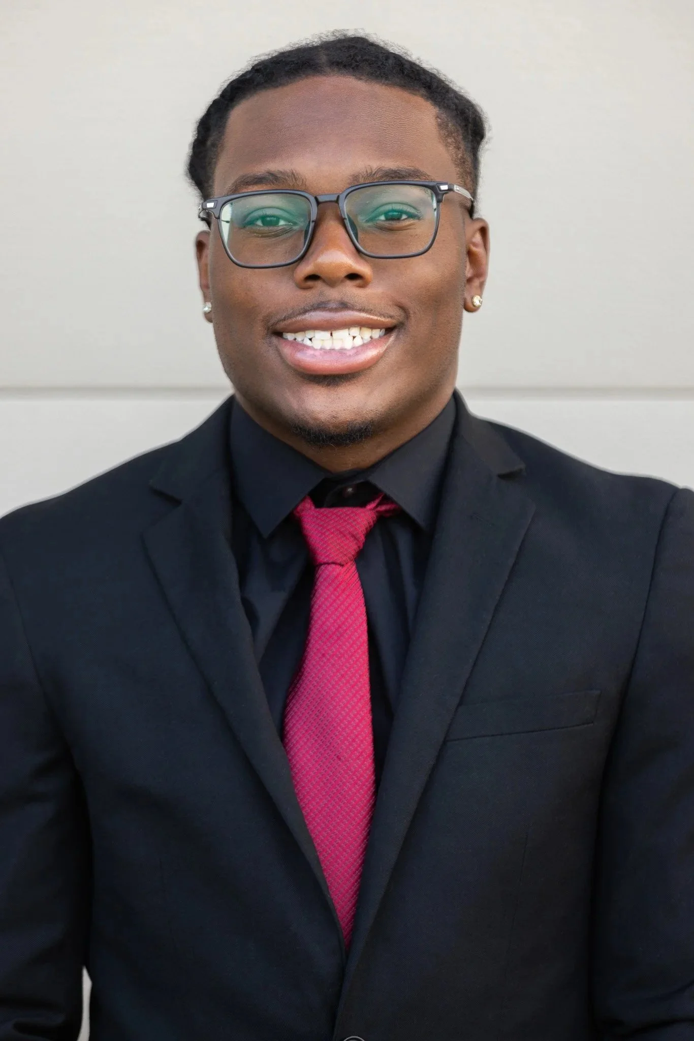 A young man with glasses, earrings, and a smiling face wearing a black suit, black shirt, and a red tie, standing against a plain light-colored background.