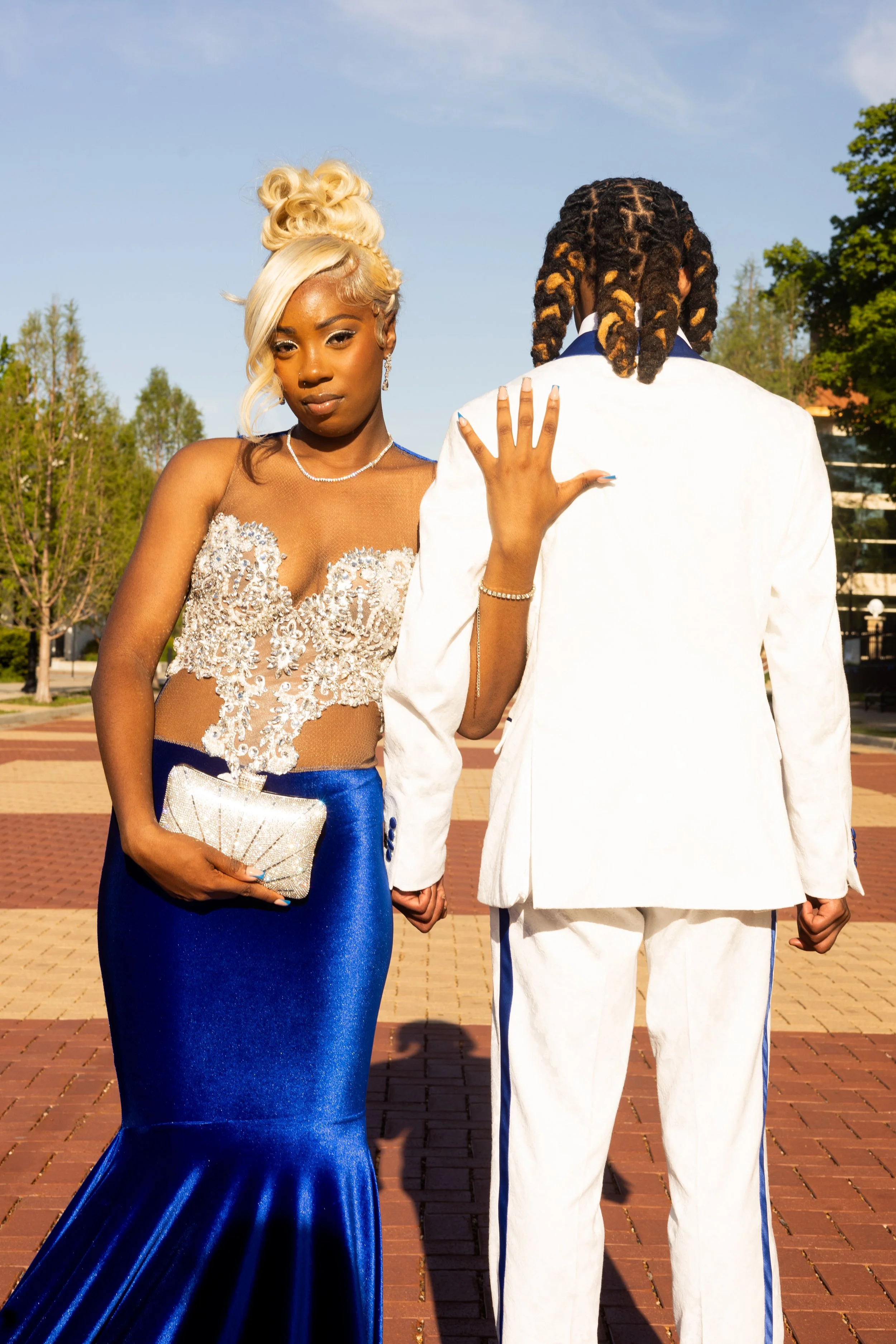 A woman in a blue gown with floral embellishments on the bodice, holding a silver clutch, and a man in a white tuxedo with blue accents, standing outdoors on a brick-paved area with trees and a building in the background.