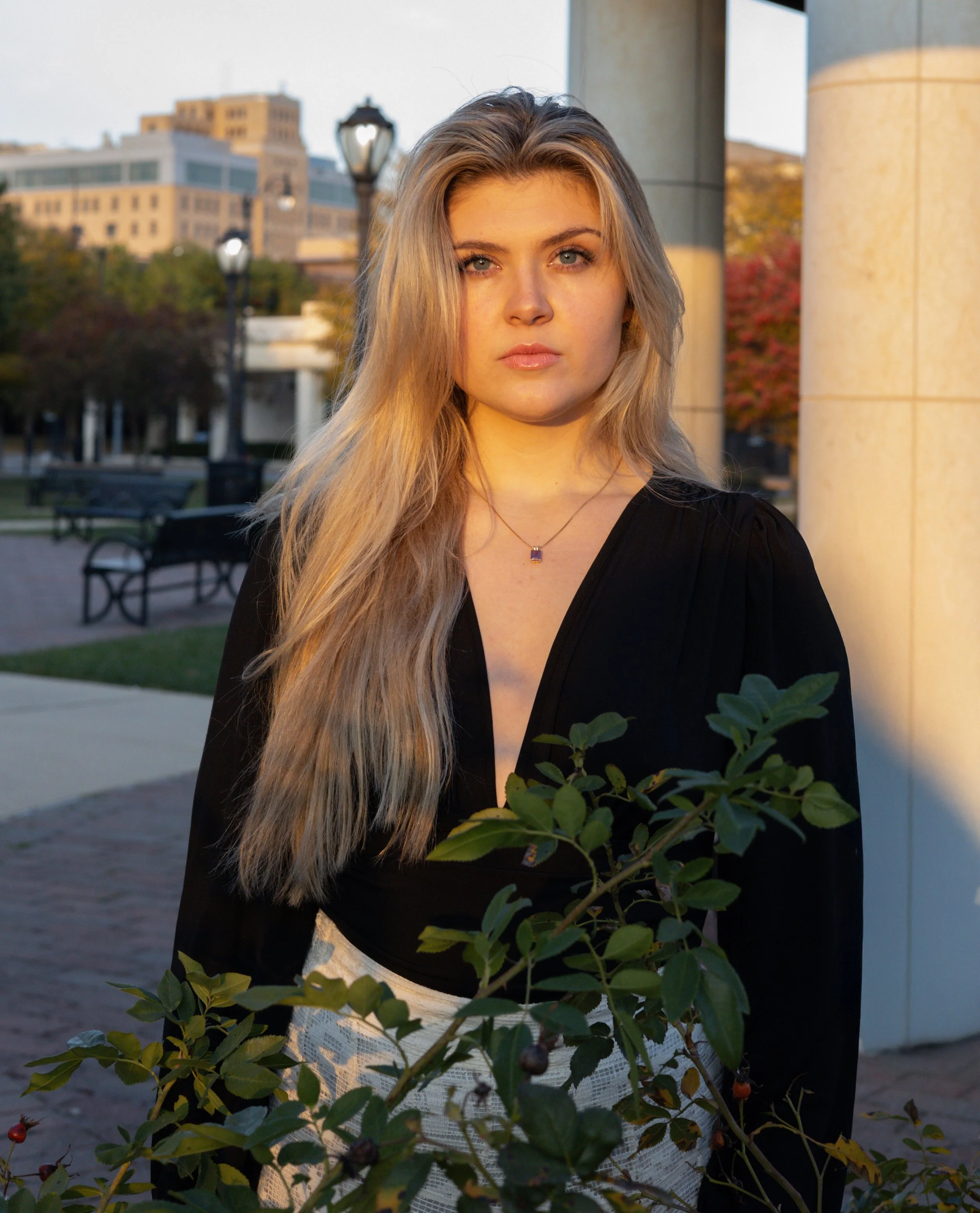 A young woman with long blonde hair and blue eyes standing outdoors during sunset, with a cityscape and trees in the background, wearing a black top and a purple gemstone necklace.